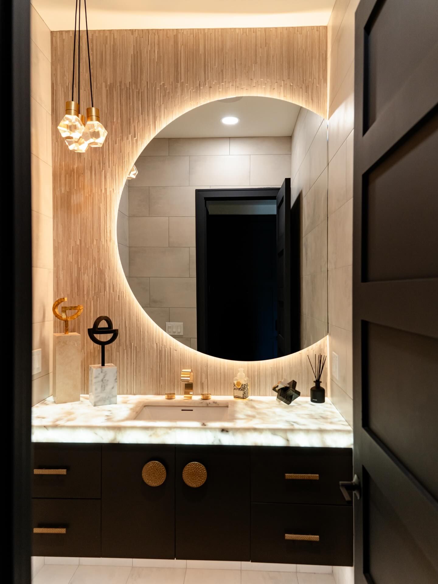 Modern bathroom vanity with round mirror, warm lighting, marble countertop, and dark cabinets