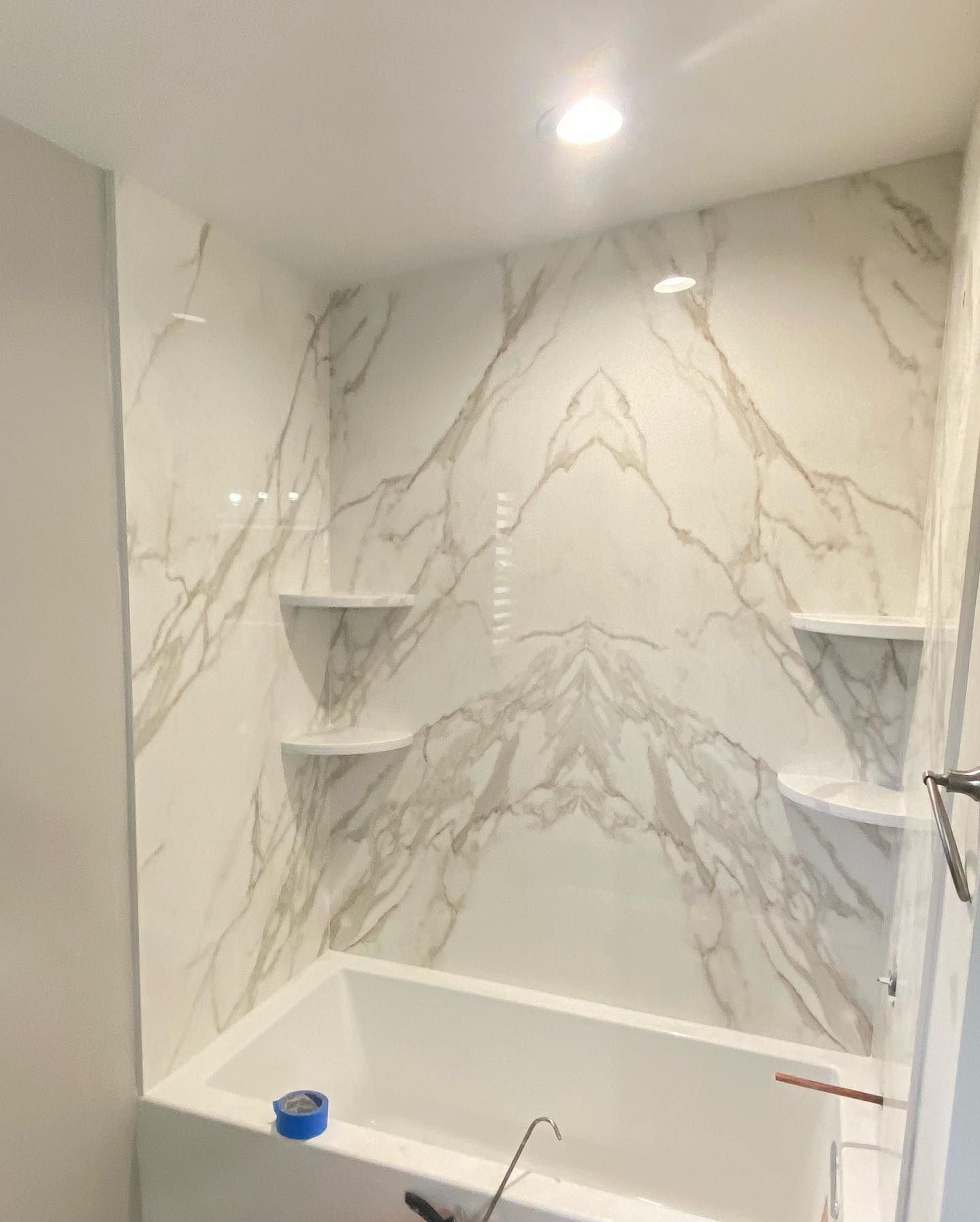 Empty marble-tiled shower with built-in corner shelves and a white bathtub ledge
