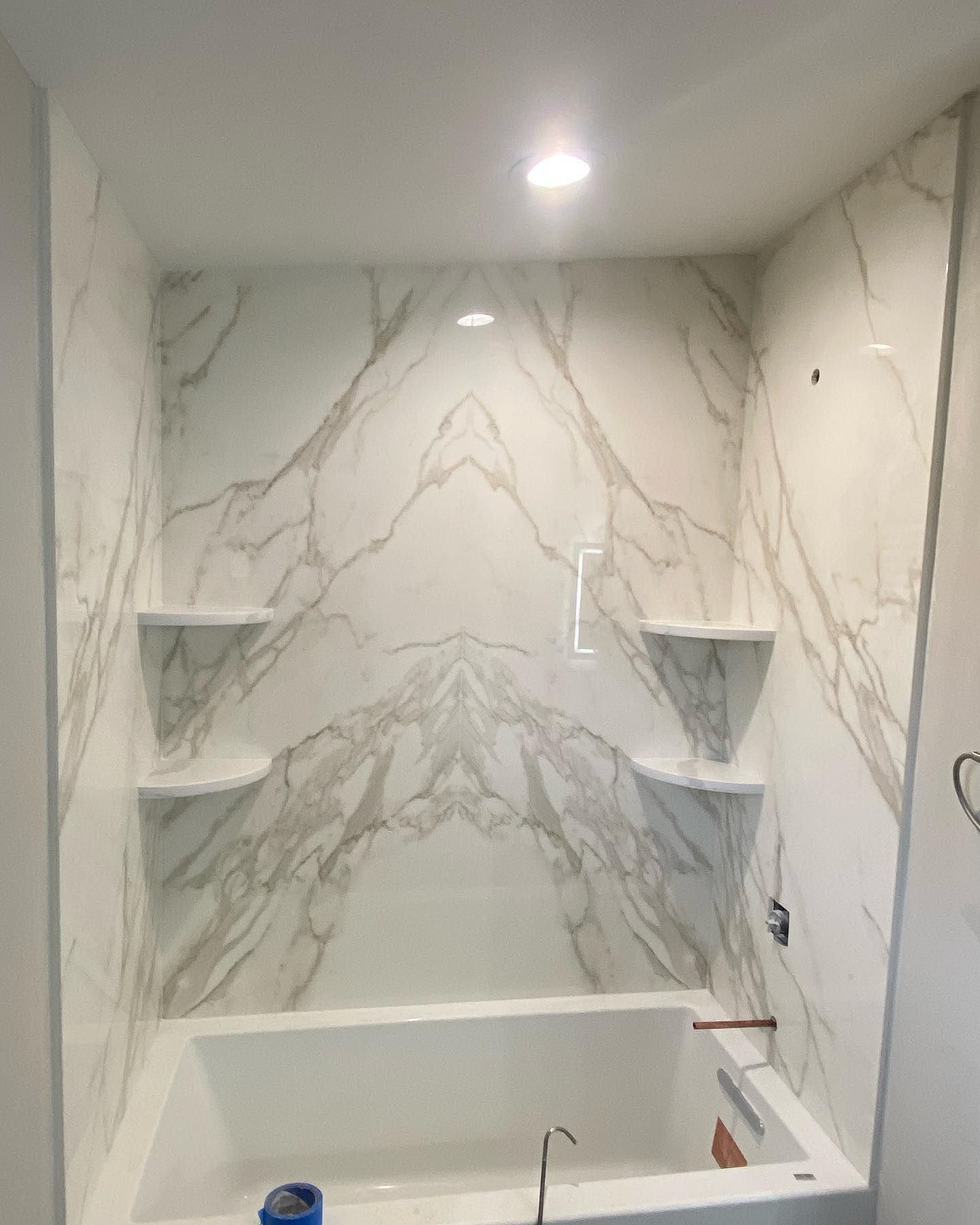 White marble-tiled shower with built-in corner shelves, a bathtub, and overhead light.