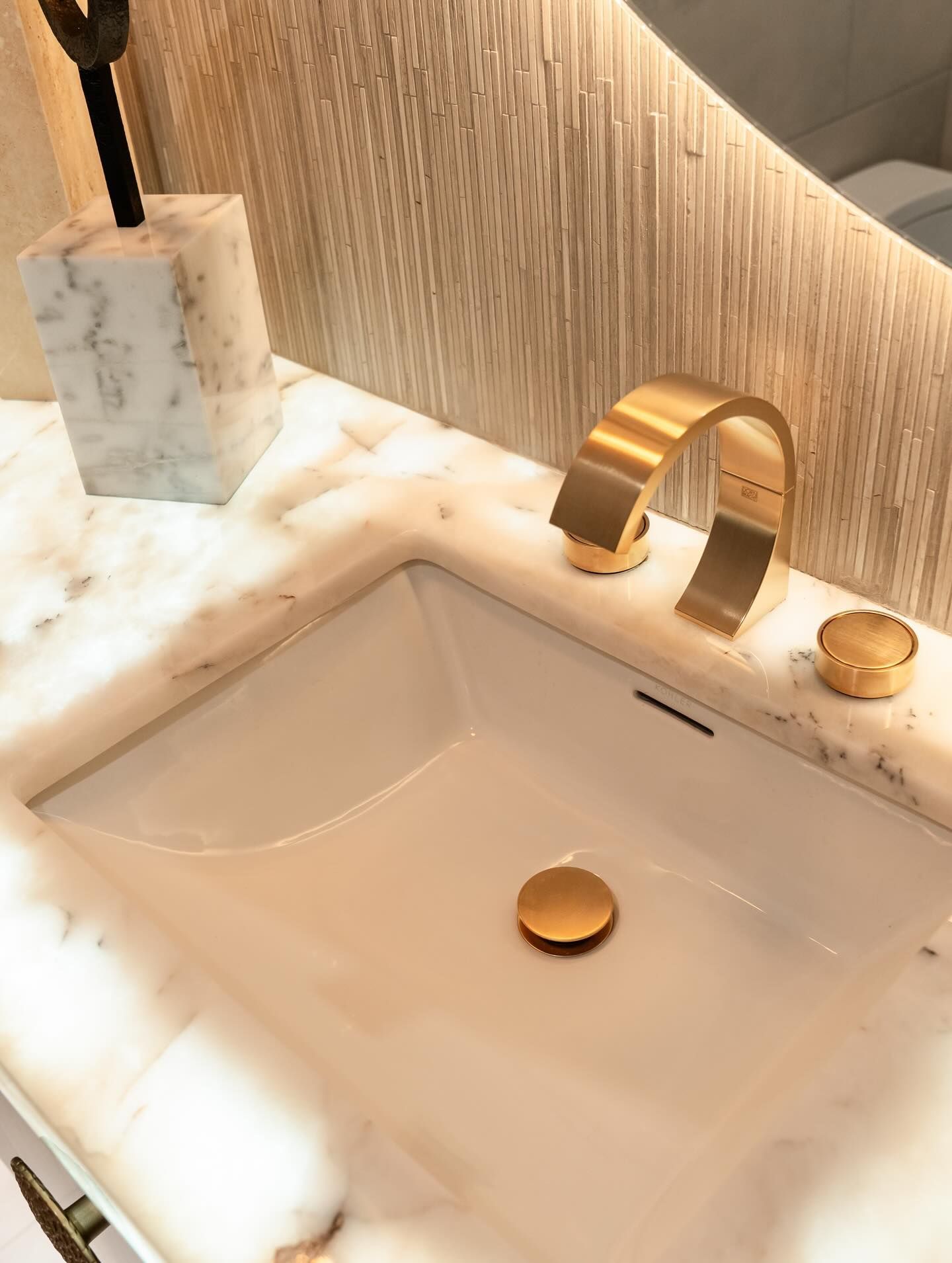 Gold faucet and drain in a white marble bathroom sink with a matching soap dispenser and knob.