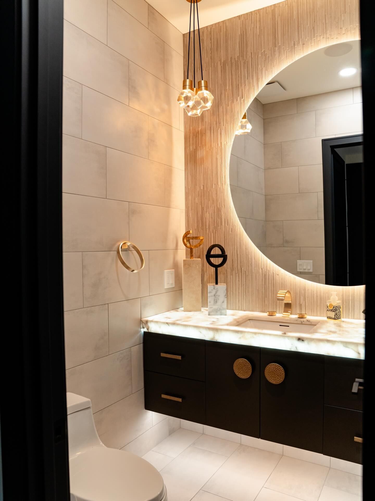 Modern bathroom vanity with round backlit mirror, dark cabinet, white tile, and gold fixtures