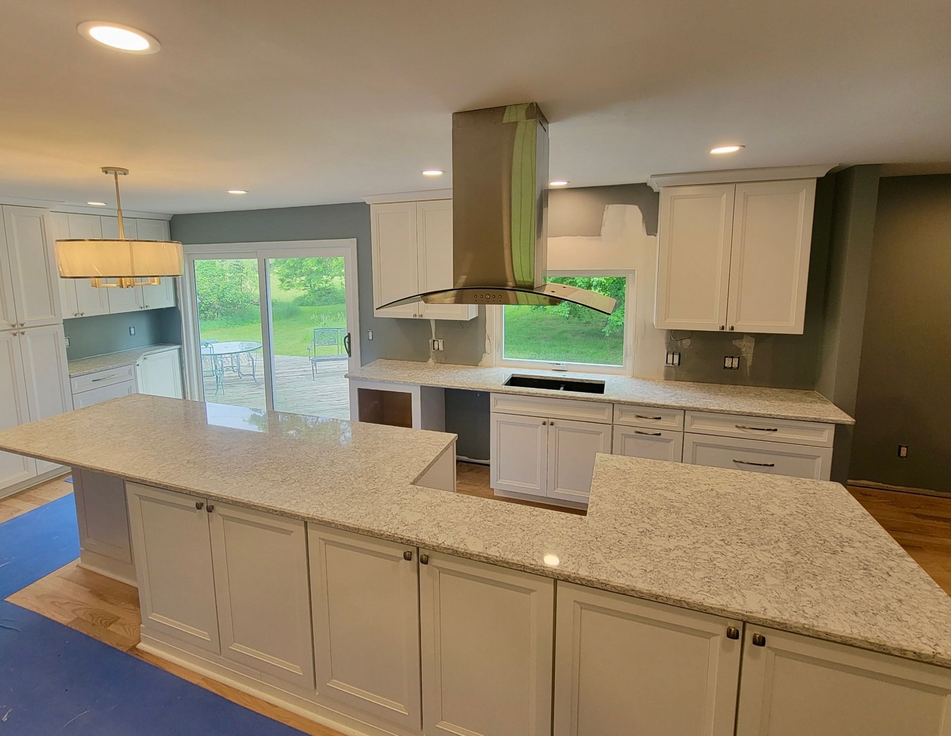 Bright kitchen with white cabinets, speckled island, stainless stove hood, and large windows overlooking greenery
