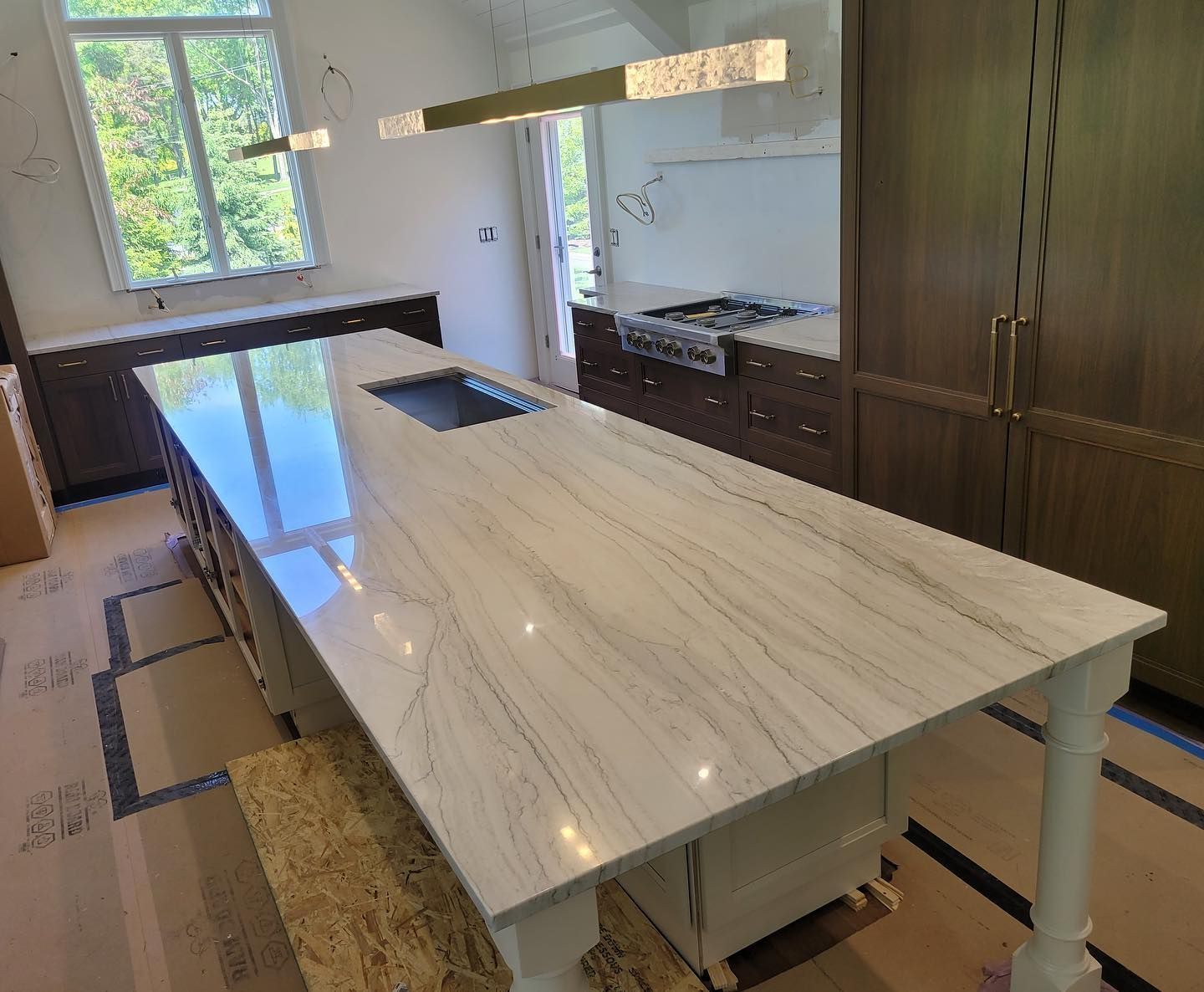 Modern kitchen with a large white marble island, dark cabinets, and a window overlooking greenery.