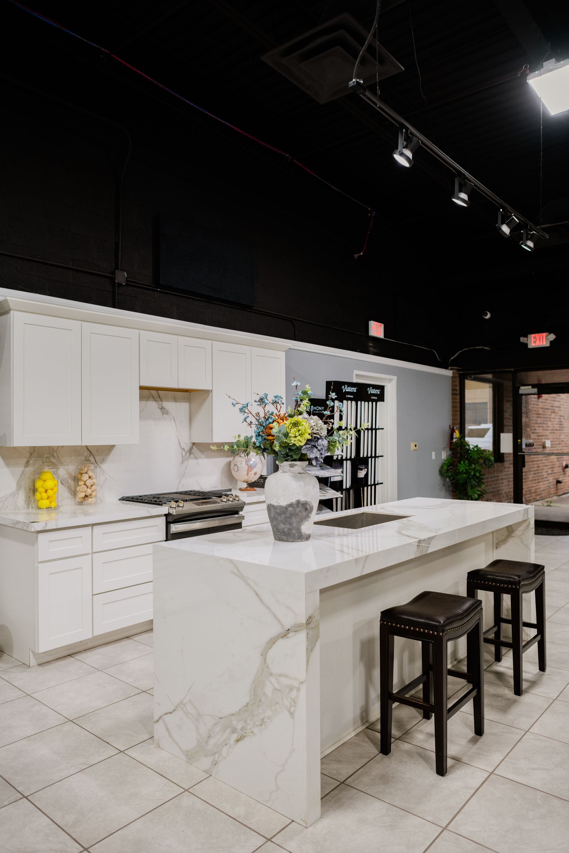 Modern white kitchen showroom with marble island, black stools, and overhead track lighting
