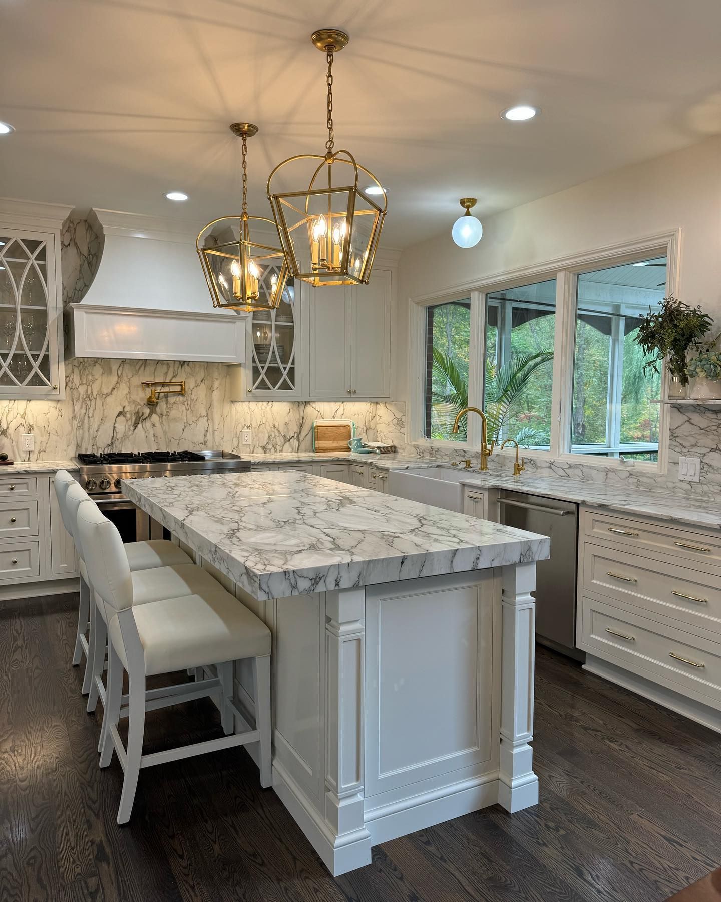 Bright white kitchen with marble island, brass pendants, and large windows overlooking greenery
