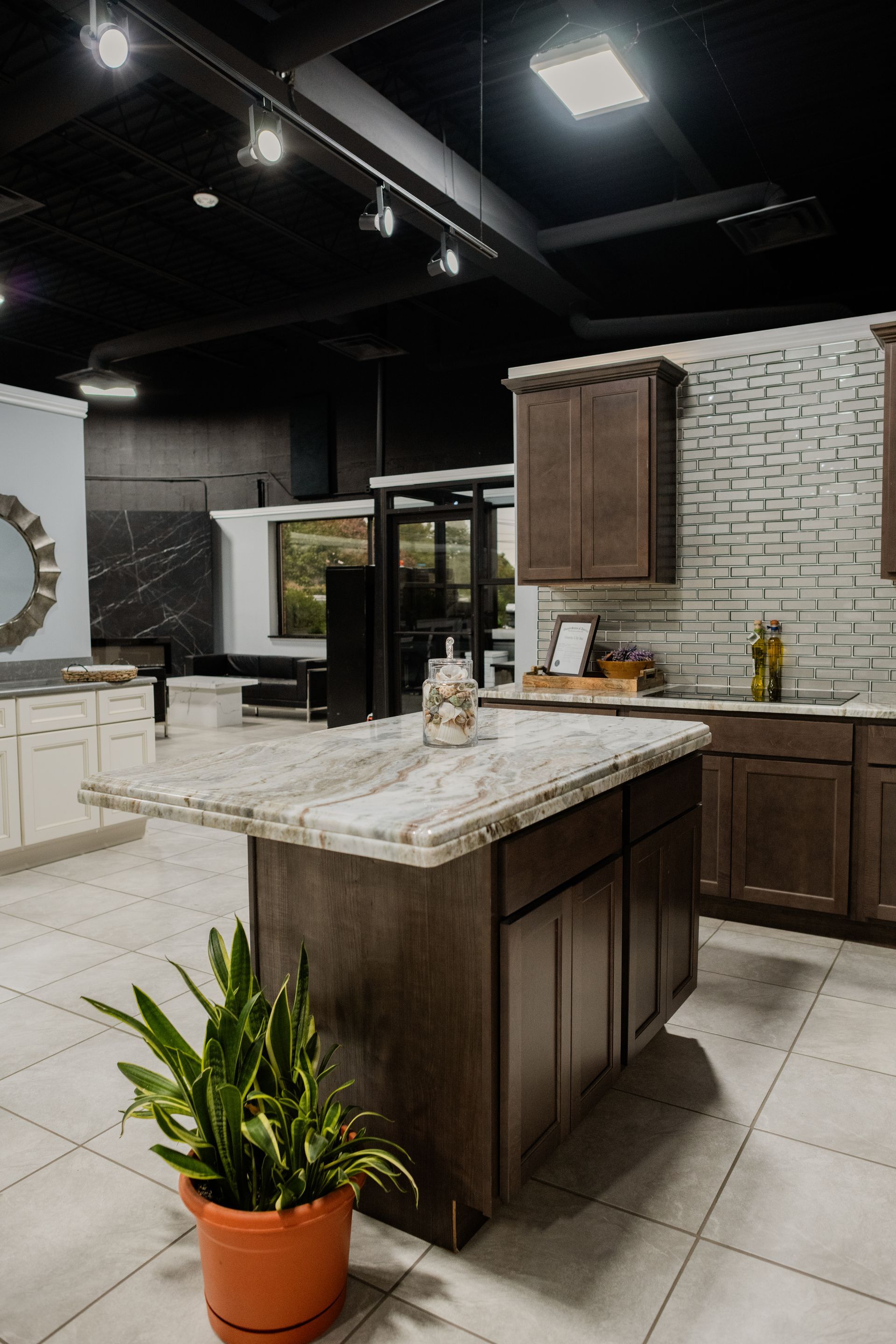 Modern kitchen showroom with dark wood cabinets, white marble island, tiled floor, and potted plant.