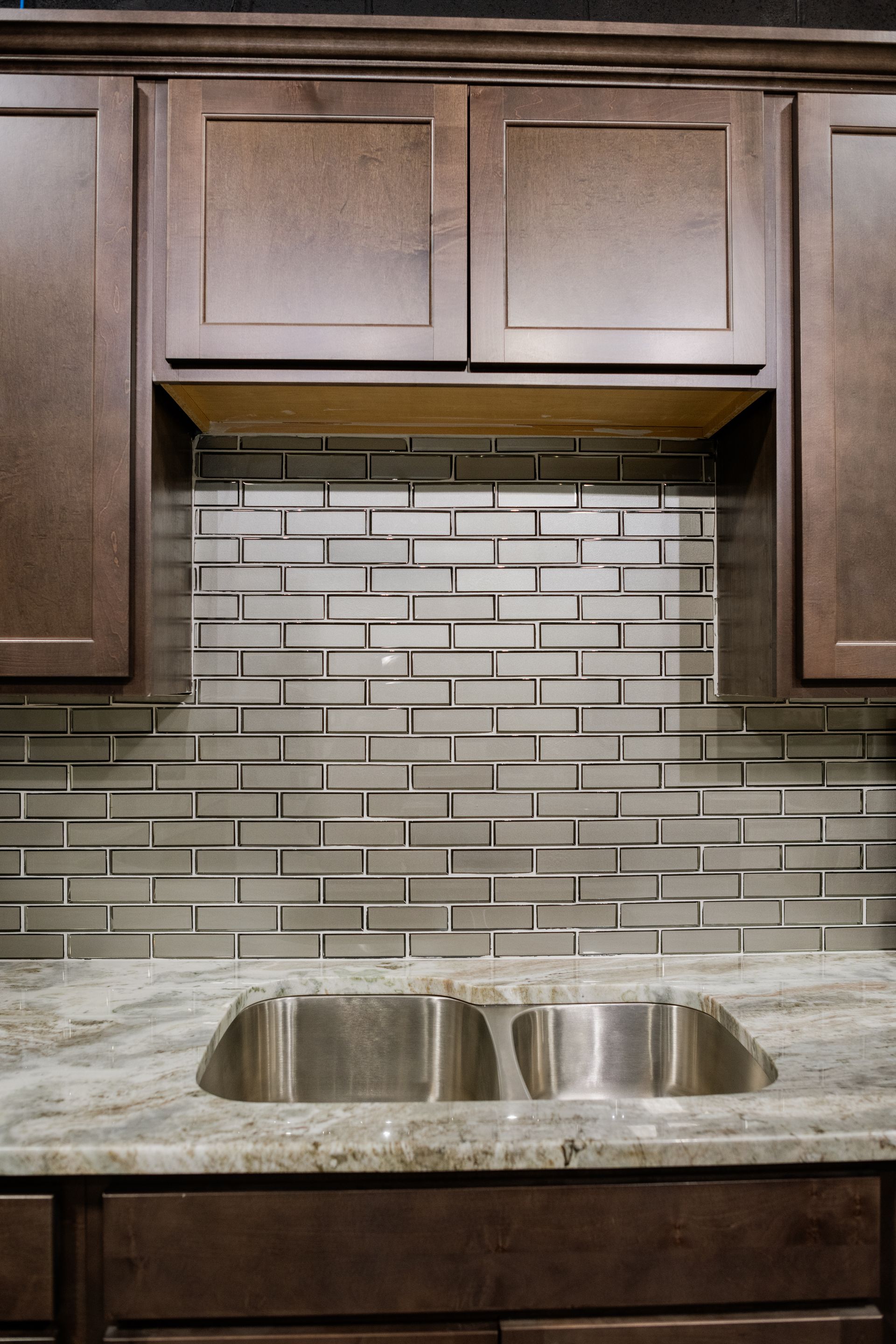 Kitchen sink with double basins, marble countertop, gray tile backsplash, and dark wood cabinets above and below