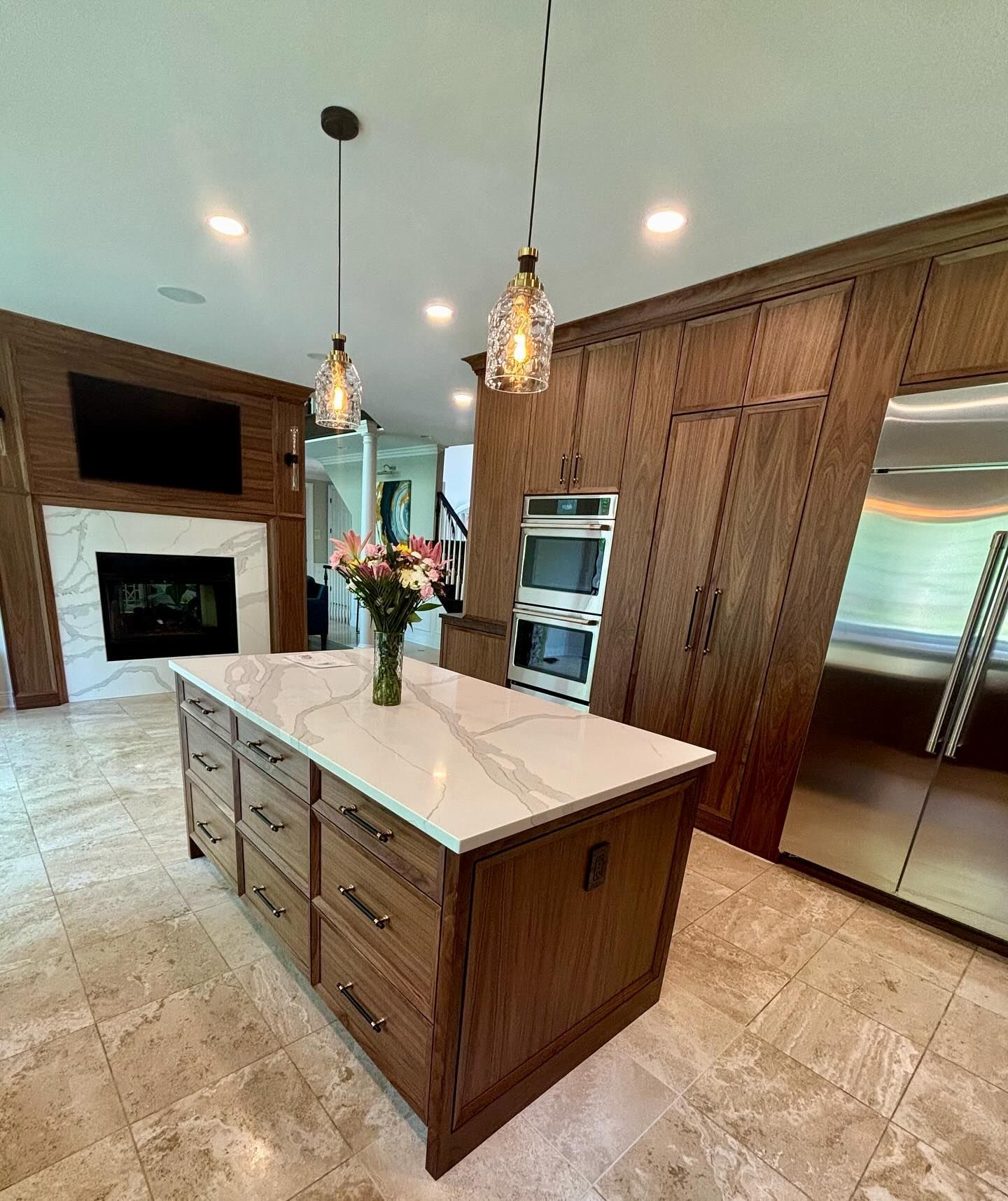 Modern kitchen with large island, white countertops, wood cabinets, pendant lights, and built-in ovens.