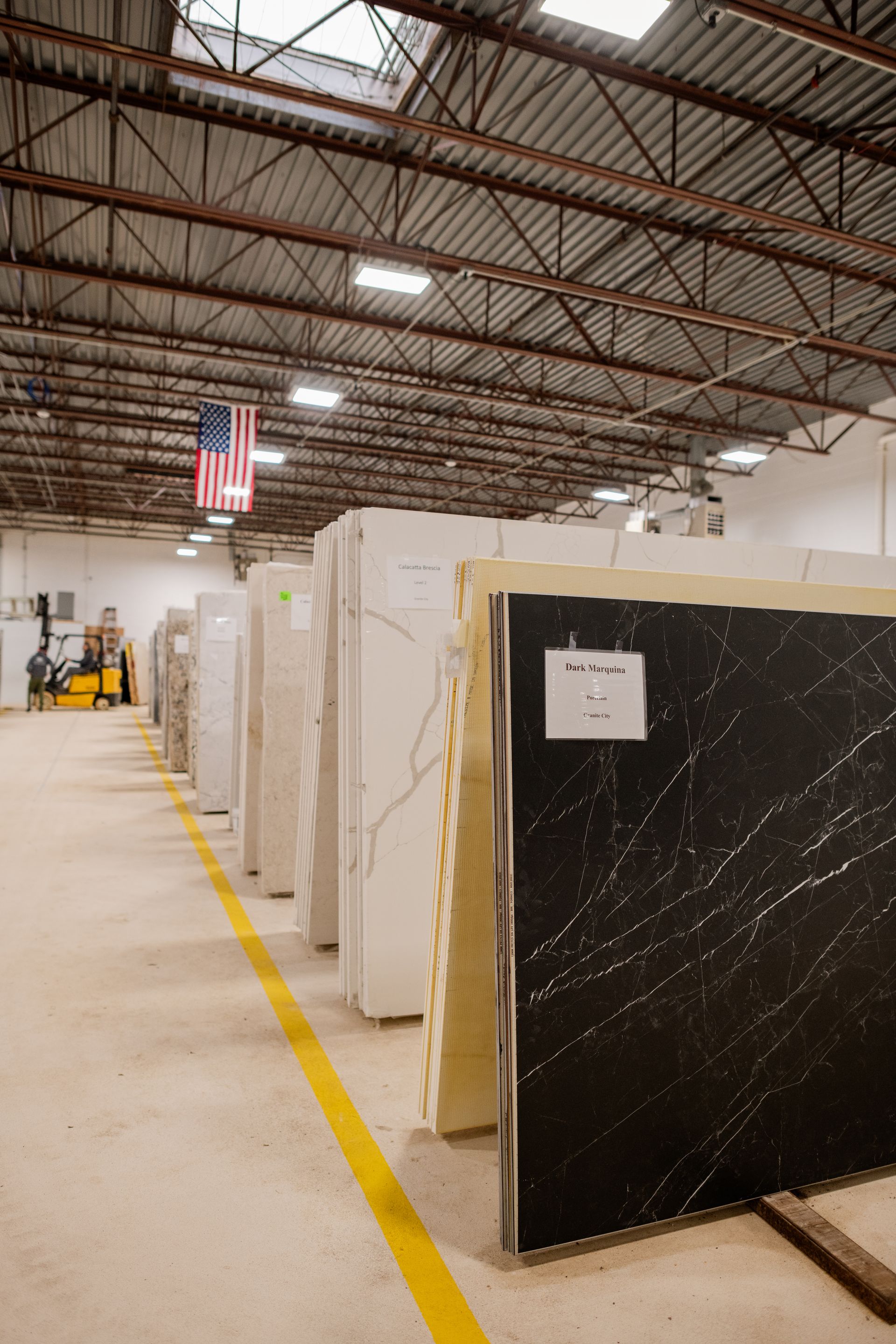 Rows of marble slabs in a warehouse aisle under high metal rafters, with a yellow floor line
