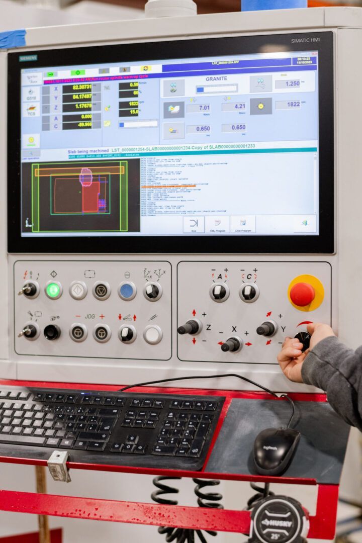 Person operating industrial control panel with monitor, buttons, keyboard, mouse, and large red emergency stop button