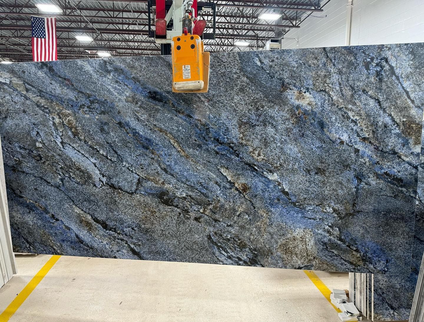 Large blue-gray granite slab suspended in an industrial warehouse by an orange crane hoist.