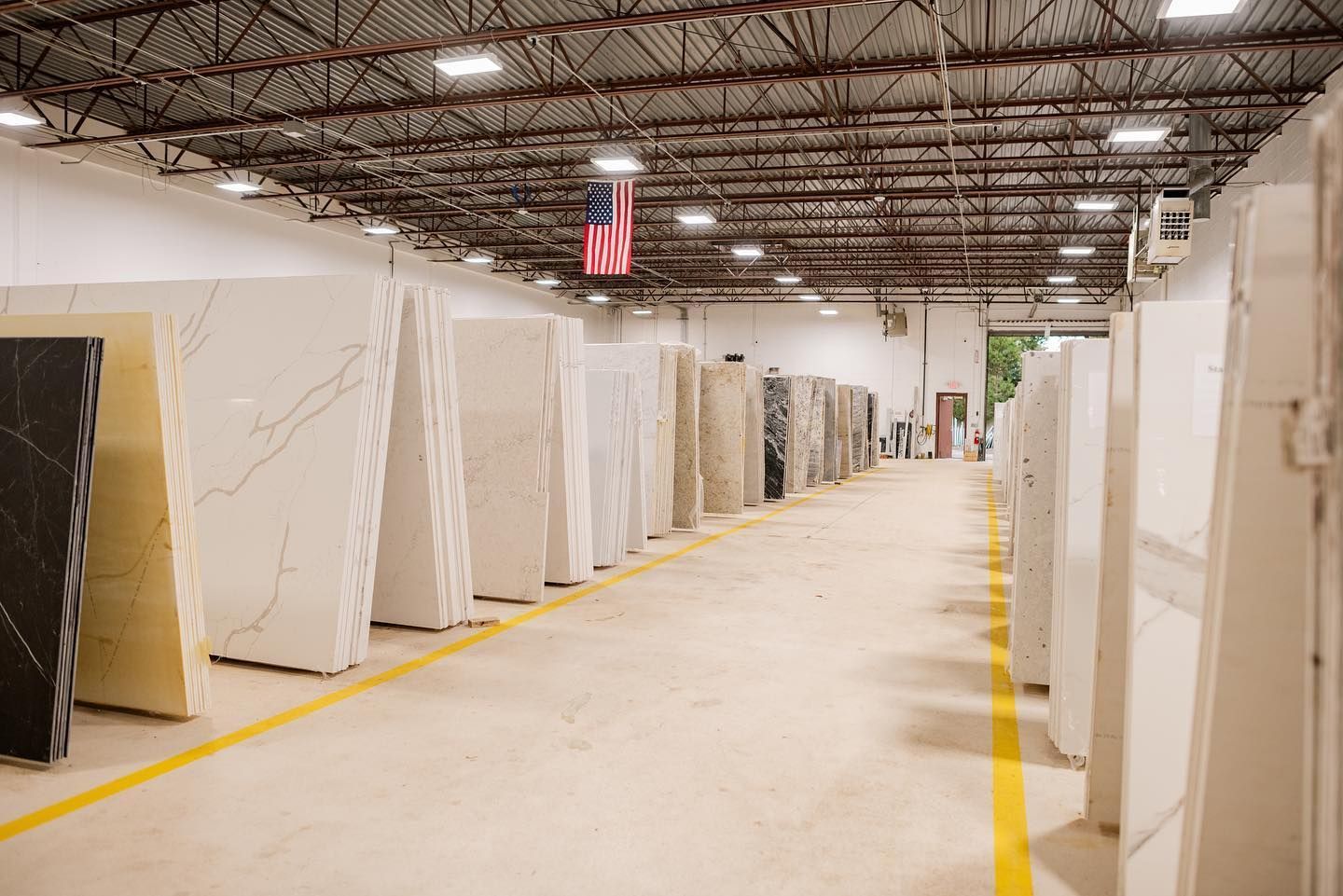 Rows of large stone slabs in a warehouse aisle under overhead lights, with an American flag hanging above.