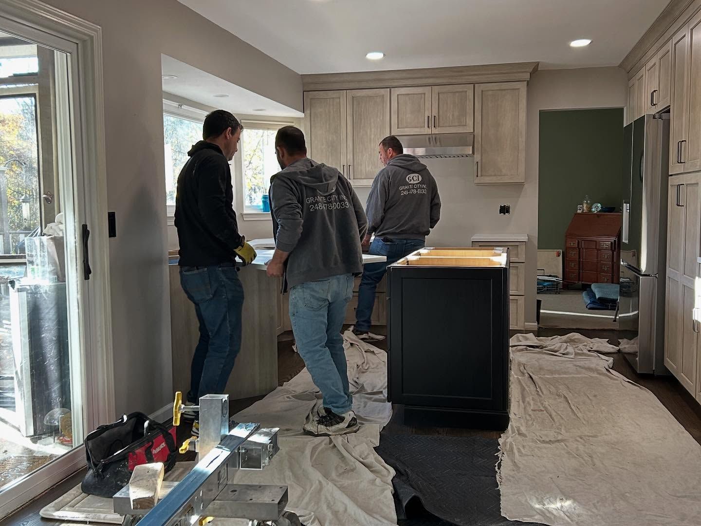 Three workers stand in an unfinished kitchen with cabinets, a center island, and tarps on the floor.