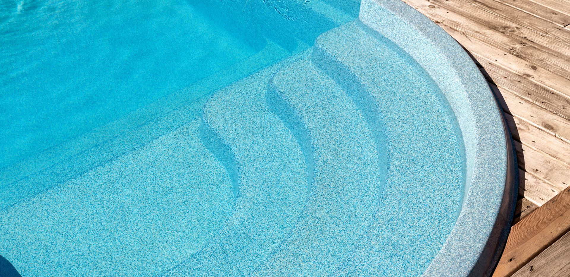 Swimming pool steps filled with bright blue water. Wooden deck on the right side.