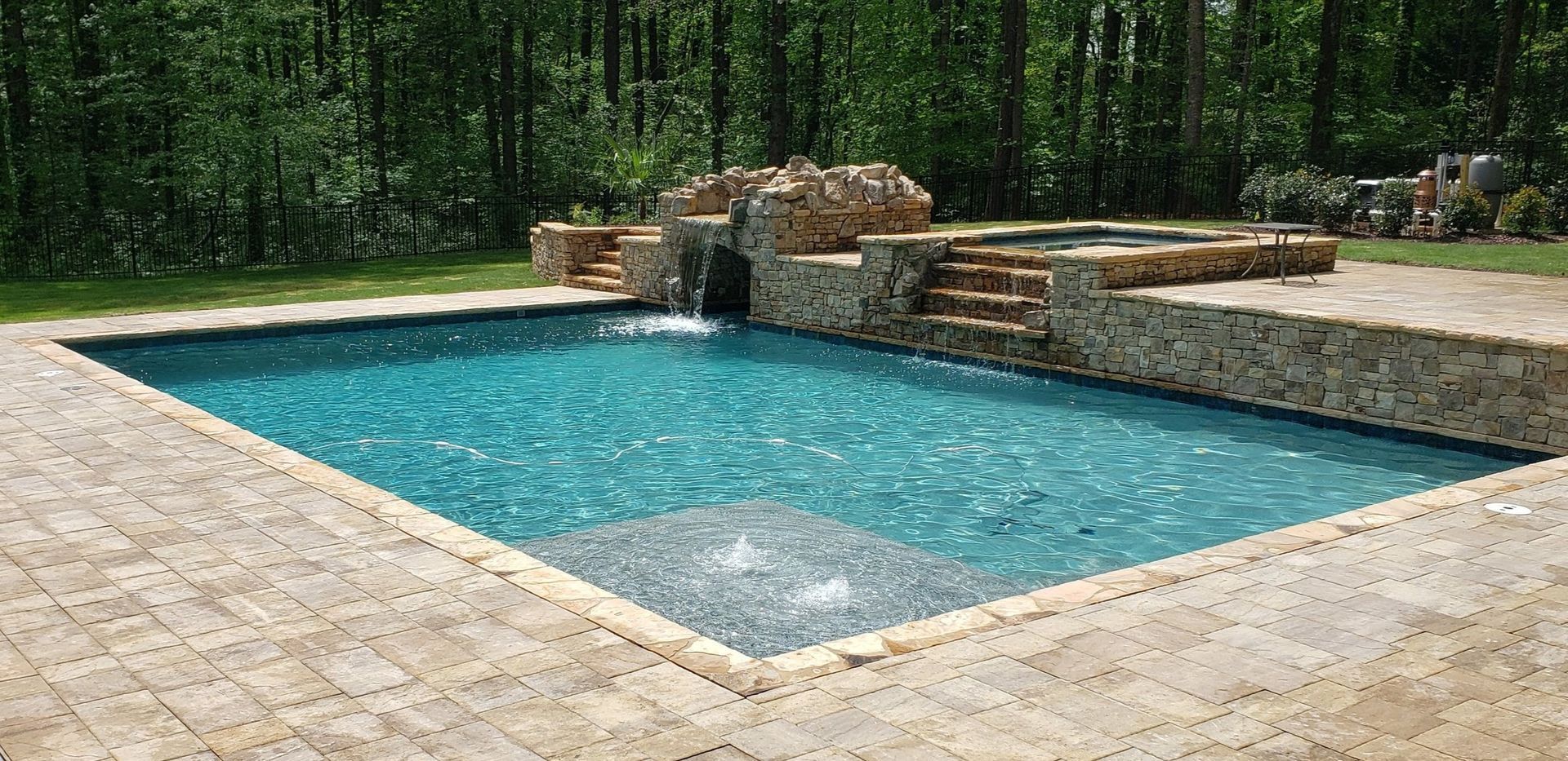 Rectangular swimming pool with a waterfall feature. Beige stone surrounds the pool. Forest in the background.