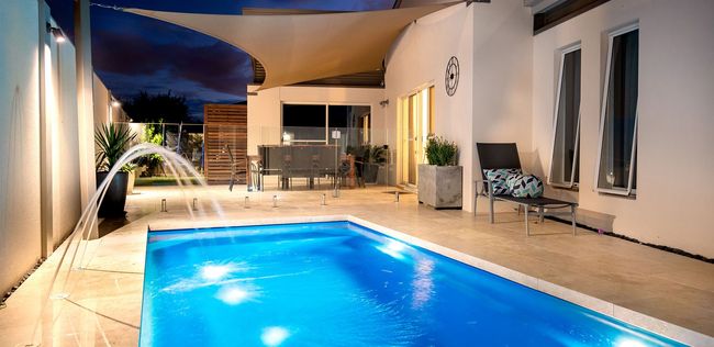 Nighttime view of a pool with a fountain, patio, and modern building with outdoor furniture.