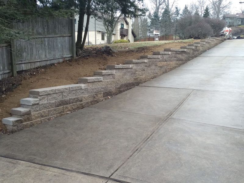 Concrete block retaining walls line a sloped driveway, partially filled with dirt. A wooden fence is to the left.
