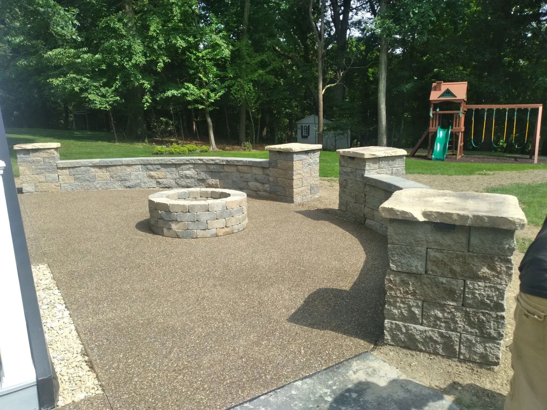 Stone patio with a fire pit, surrounding stone seating, and a playground in the background.