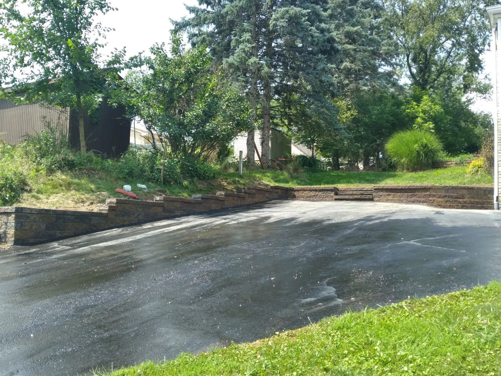 Asphalt driveway leading to a sloped yard with retaining walls and steps, trees and shrubs in the background.