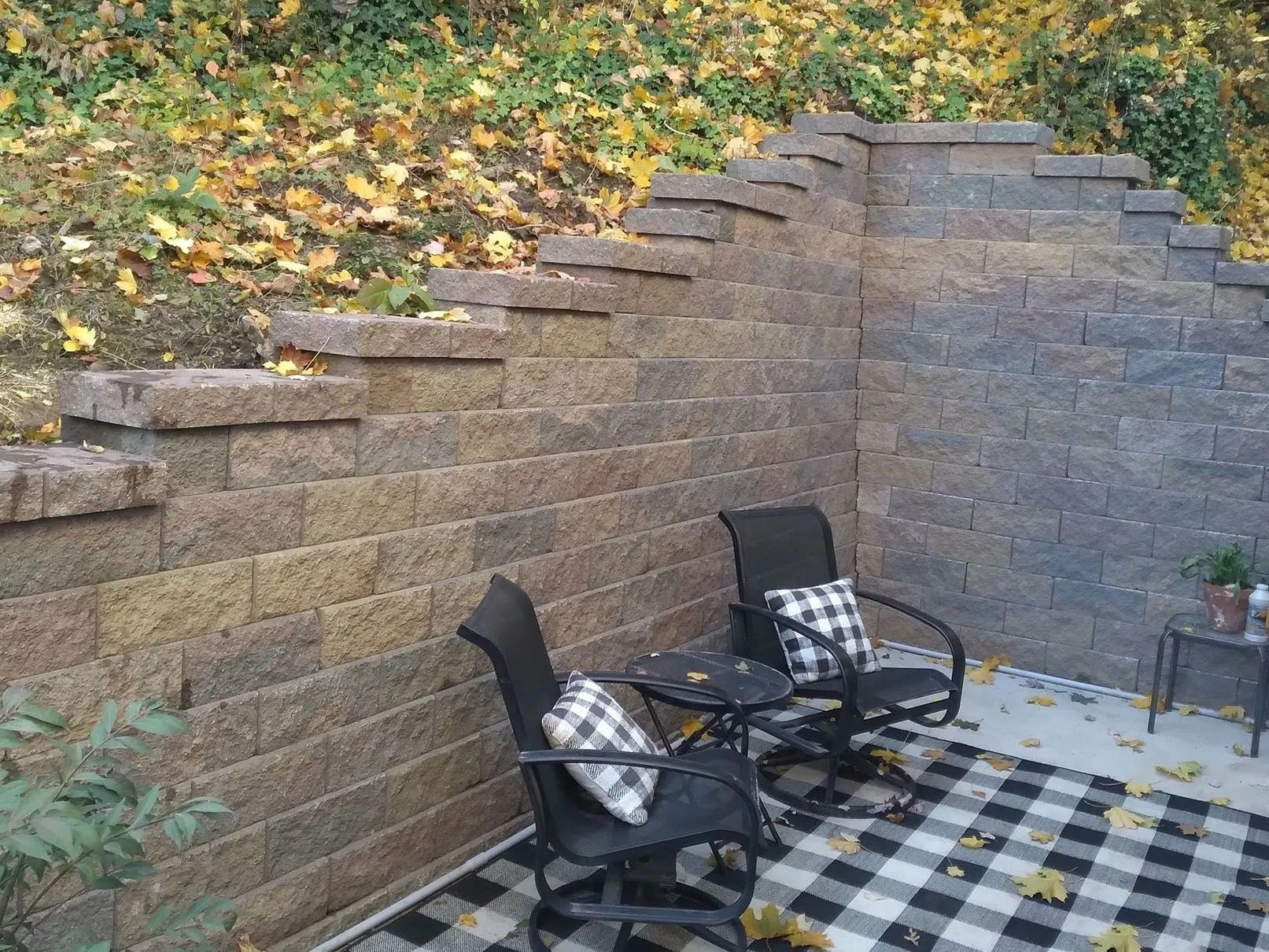 Patio seating area with stone retaining walls and fallen leaves. Two black chairs with plaid pillows surround a small table, set on a plaid rug.