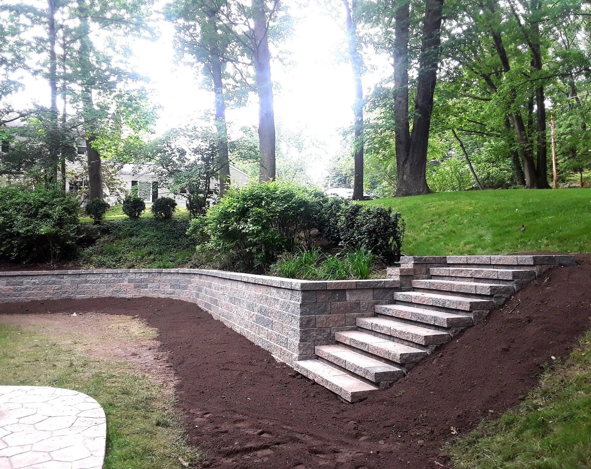Stone steps and retaining wall lead up a grassy slope in a residential yard. Brown mulch borders the stairs and wall.