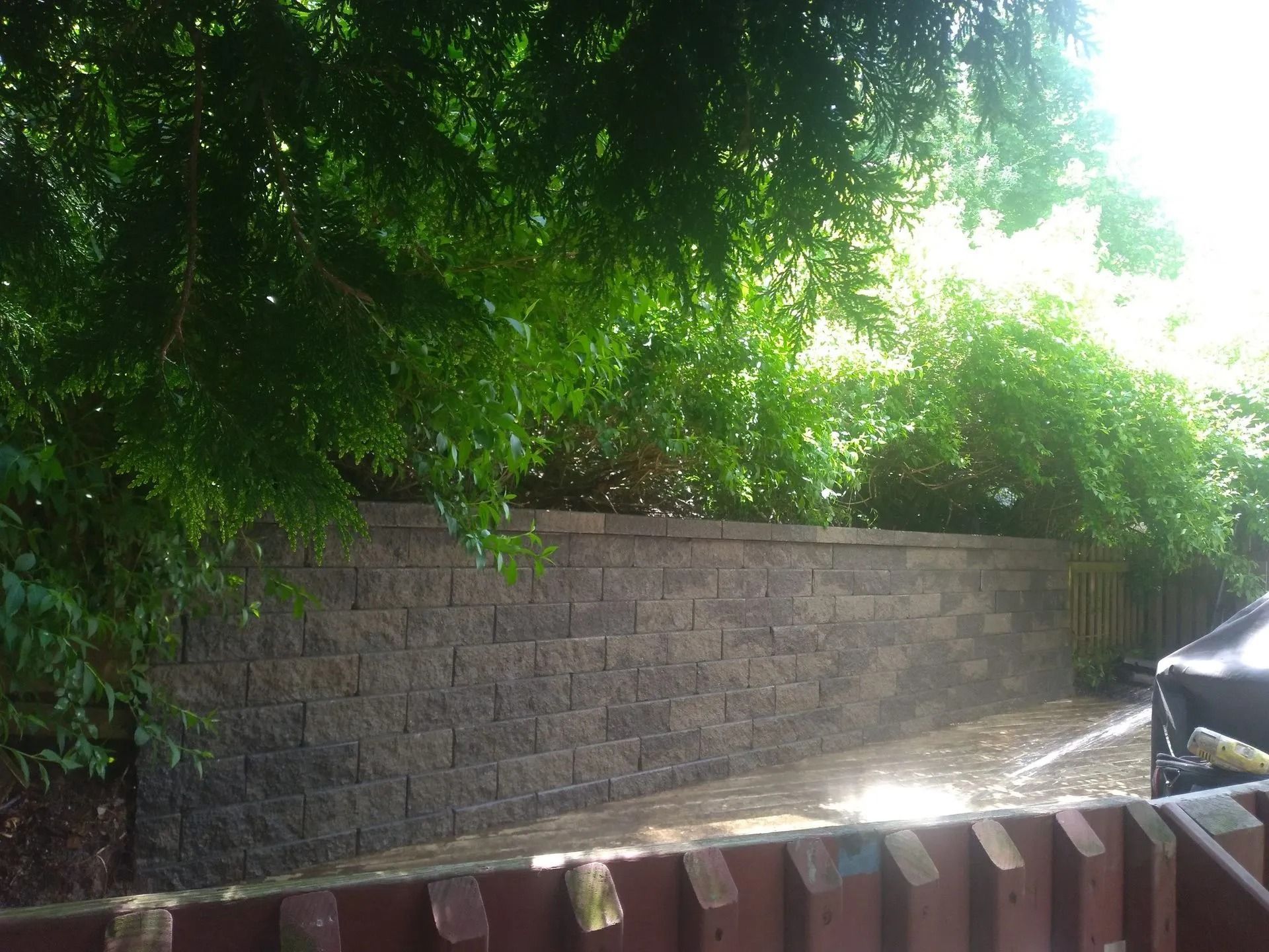 A dark gray brick retaining wall in front of lush green foliage, with a paved area and the edge of a wooden fence visible.