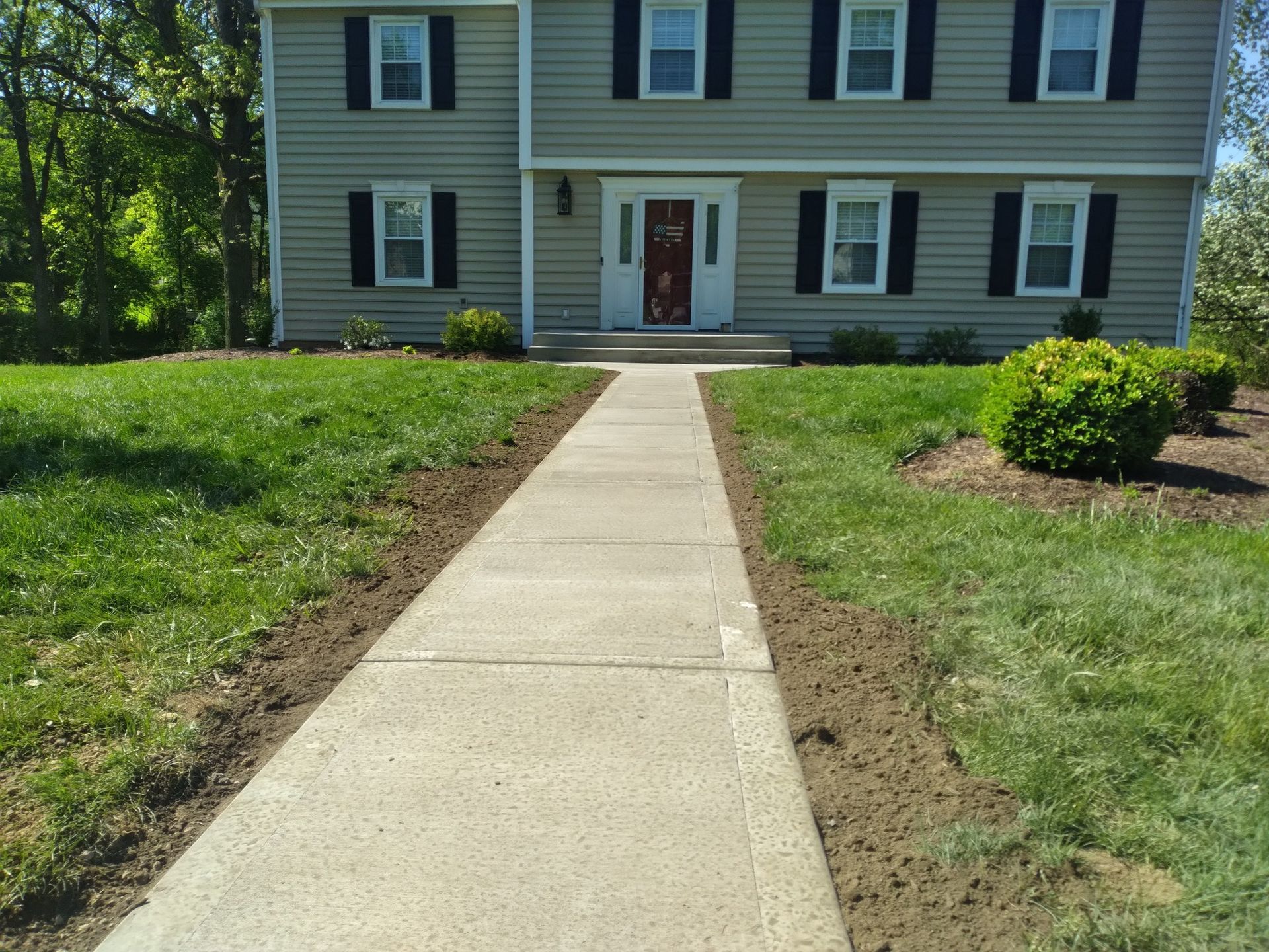 Concrete walkway leading to a two-story house with green siding and black shutters. The lawn on either side is freshly edged.
