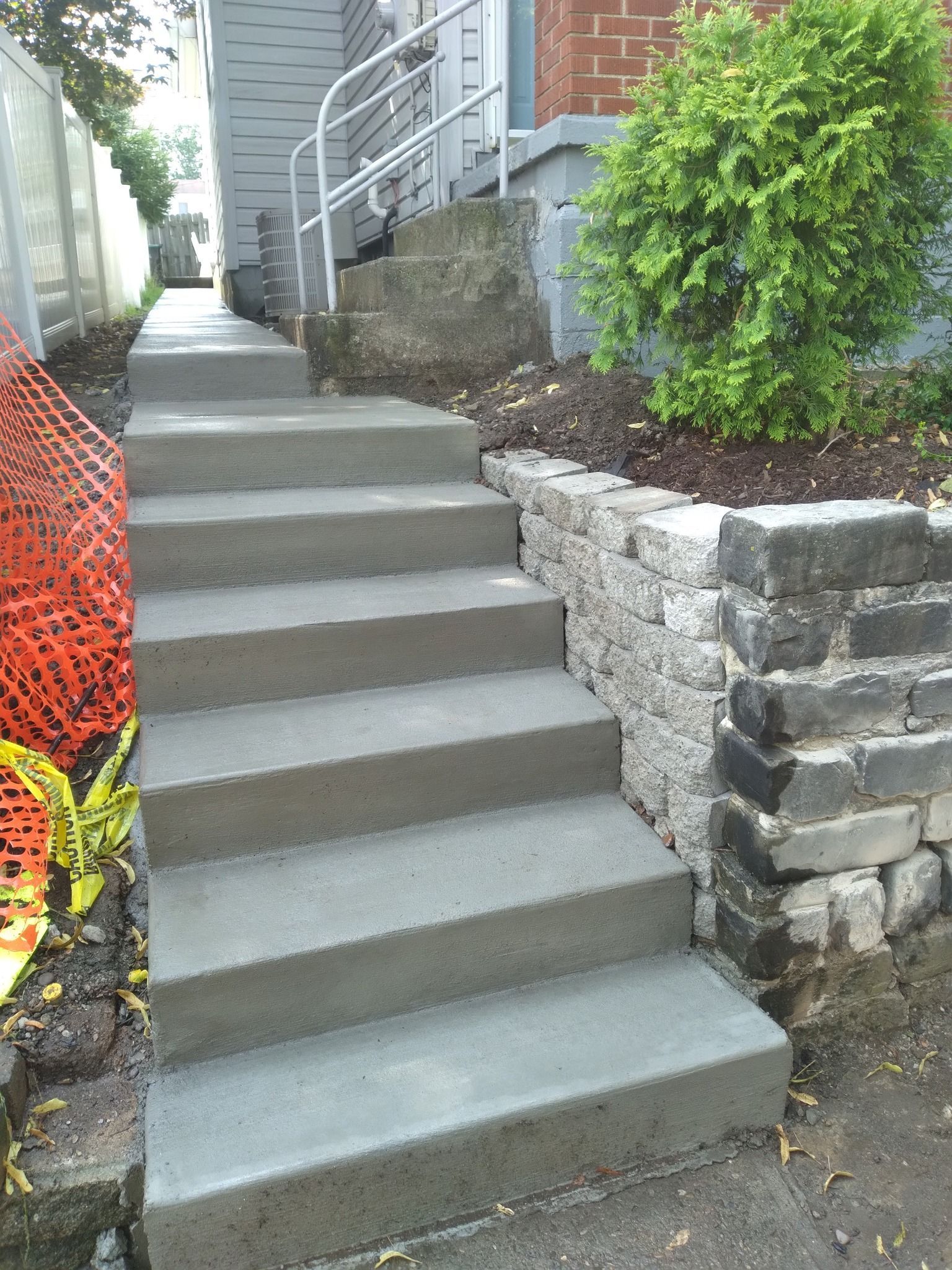 Concrete steps leading up to a building, with a handrail. Red safety fencing and a brick retaining wall are visible.