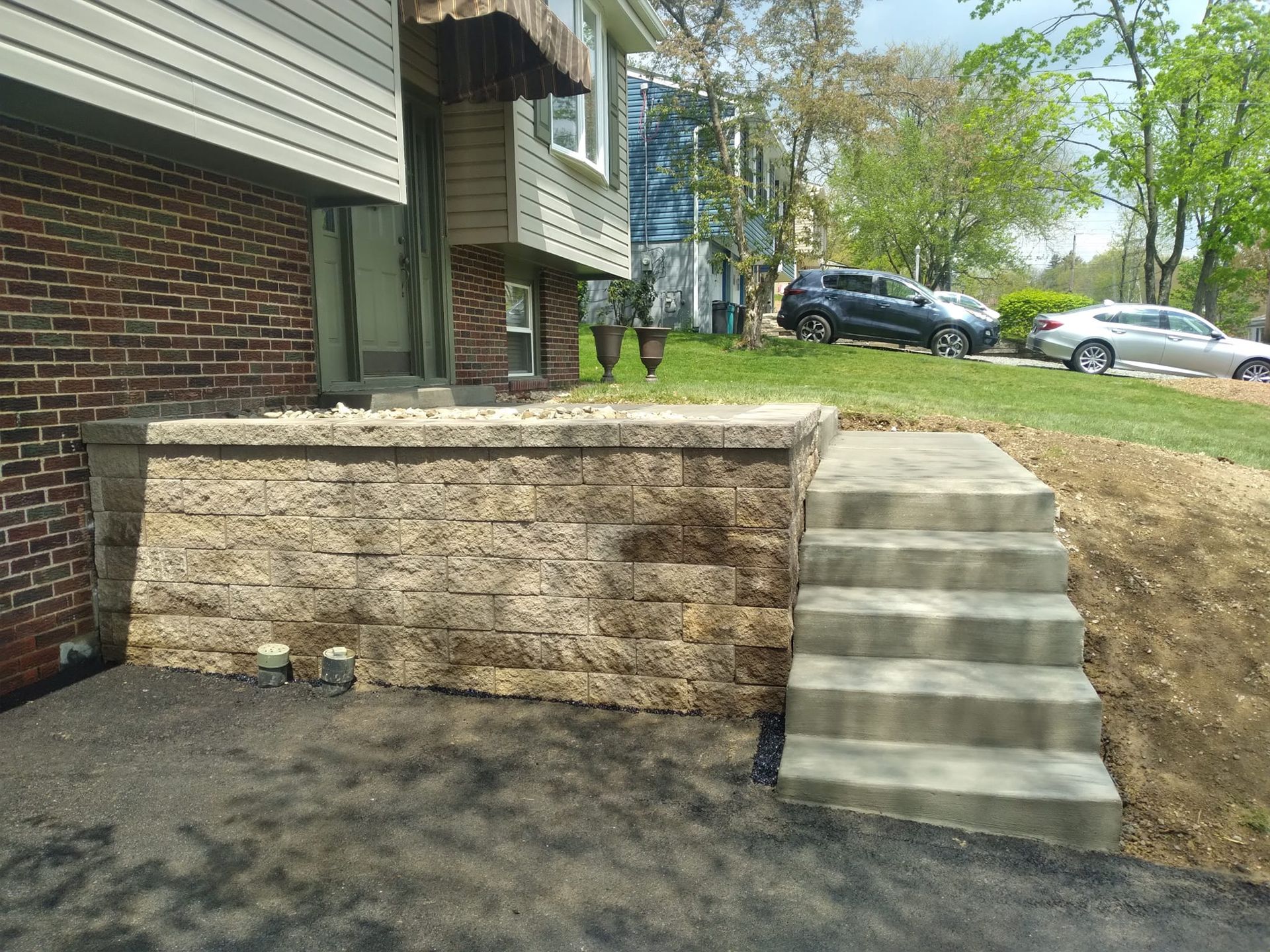 A brick house with a concrete block retaining wall and steps leading to the front door. Cars are parked in the background.