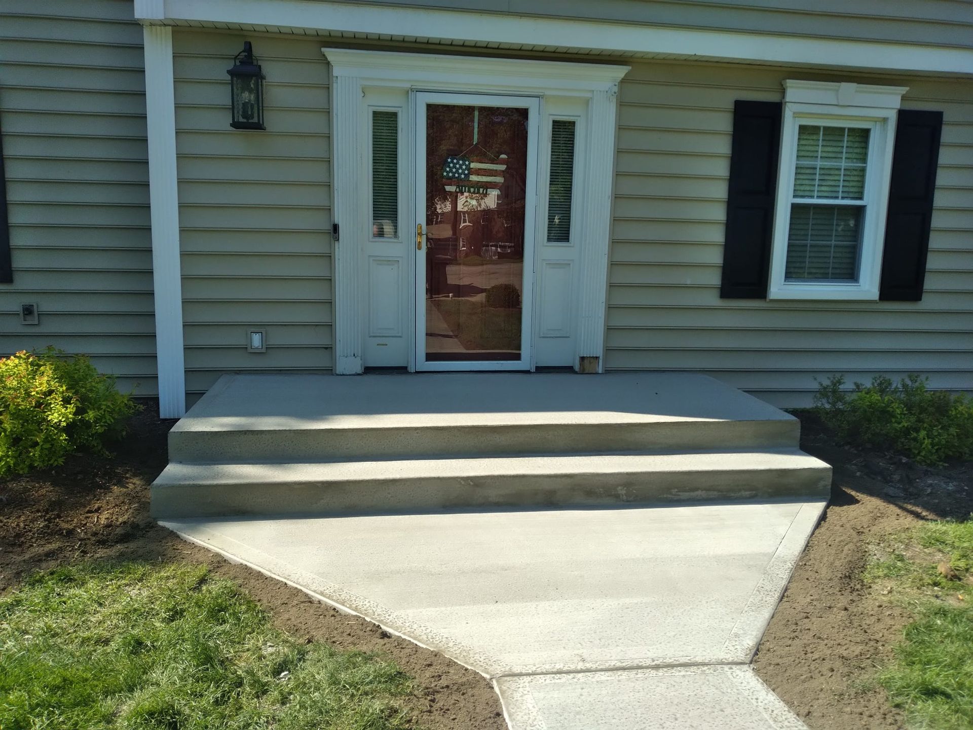Concrete steps leading up to a front door of a house with light siding and a window with black shutters.