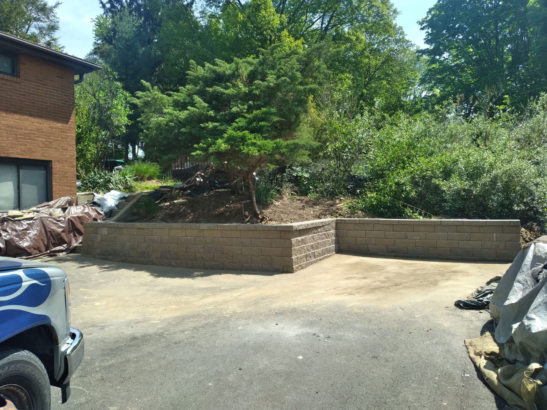 A tan brick retaining wall surrounds a small parking area. A small tree grows in the center, with trees and a house in the background.
