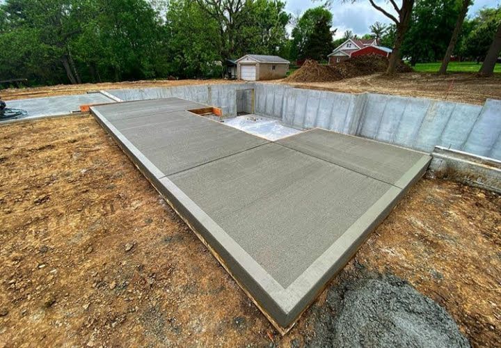 Newly poured concrete foundation, L-shaped, surrounded by dirt and foundation walls; small shed visible in the background.