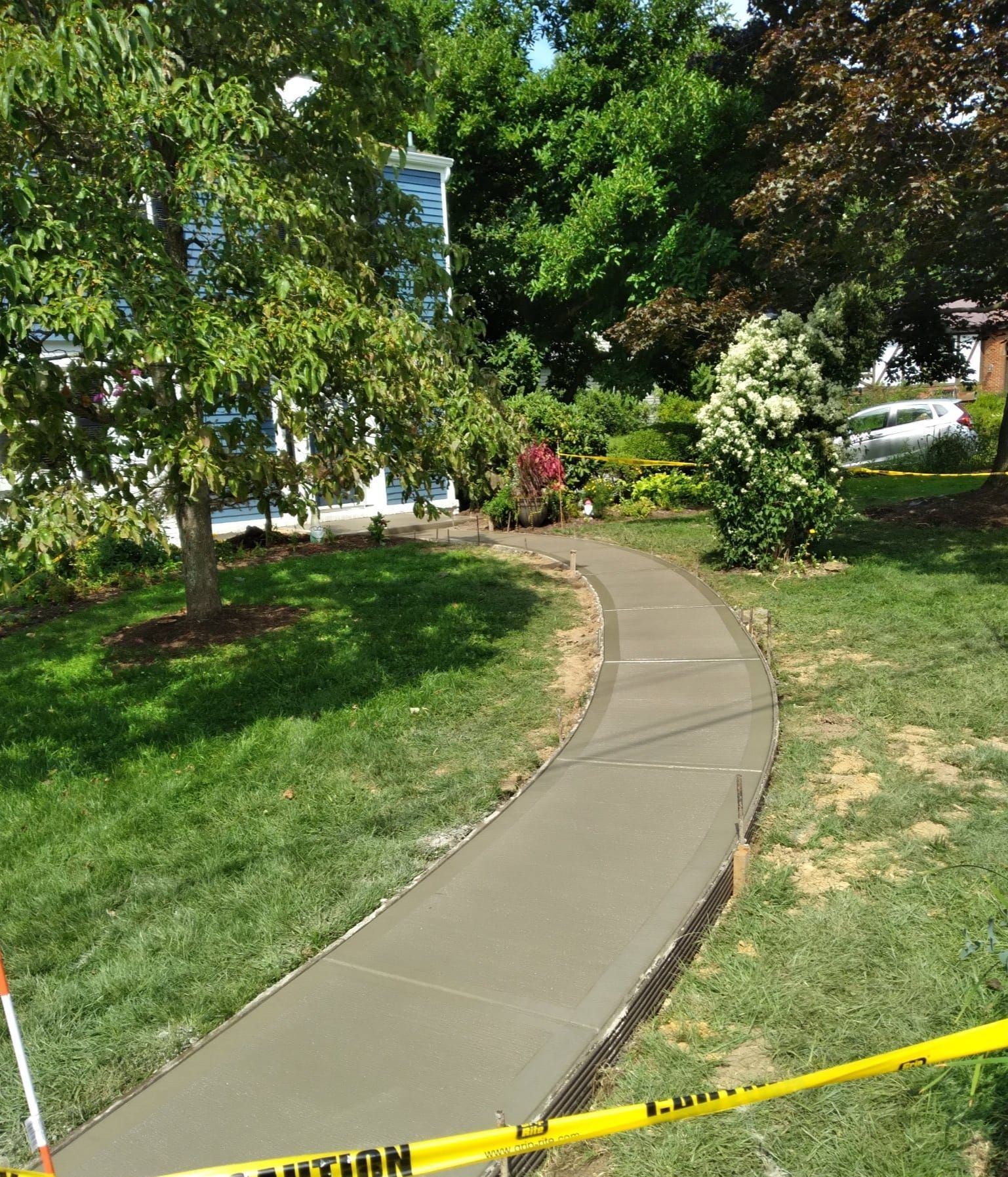 A newly poured concrete walkway curves through a grassy yard, with yellow caution tape in the foreground, leading toward a house.