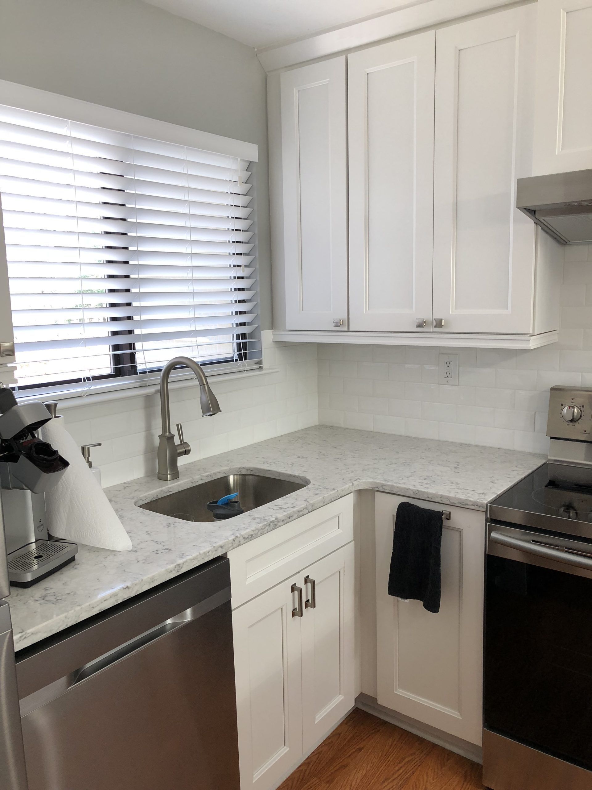 Bright white kitchen with sink, stainless dishwasher, stove, and window blinds beside a counter coffee maker