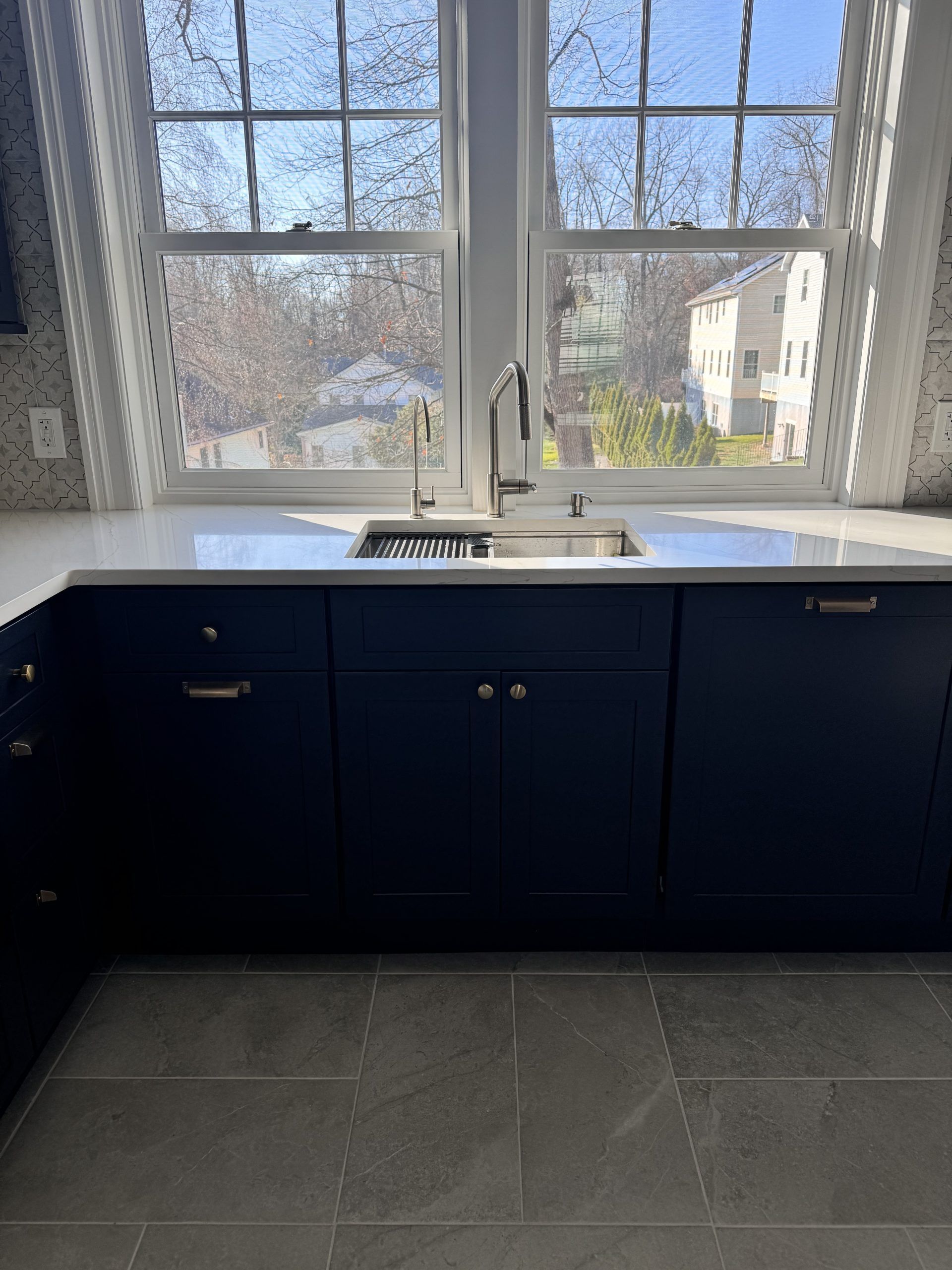 Modern kitchen sink under large windows with dark cabinets and gray tile floor