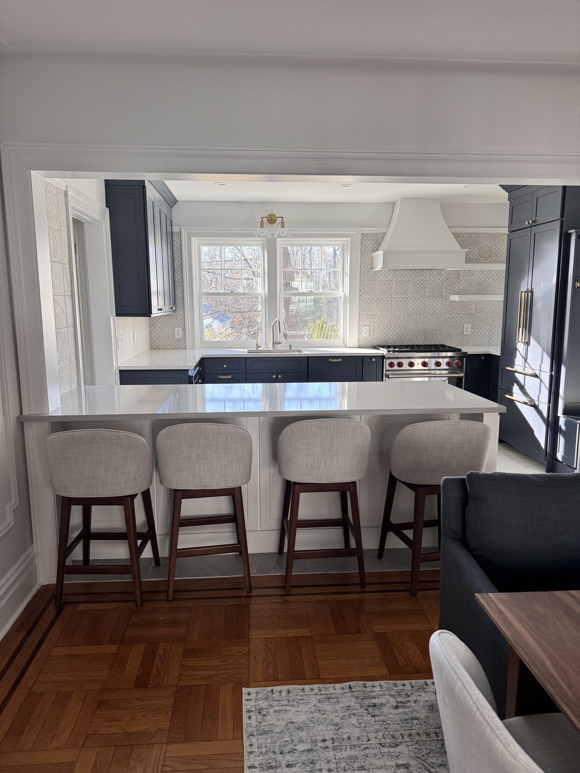 Modern kitchen seen through an open archway, with four bar stools at a white island and a dark sofa in the foreground
