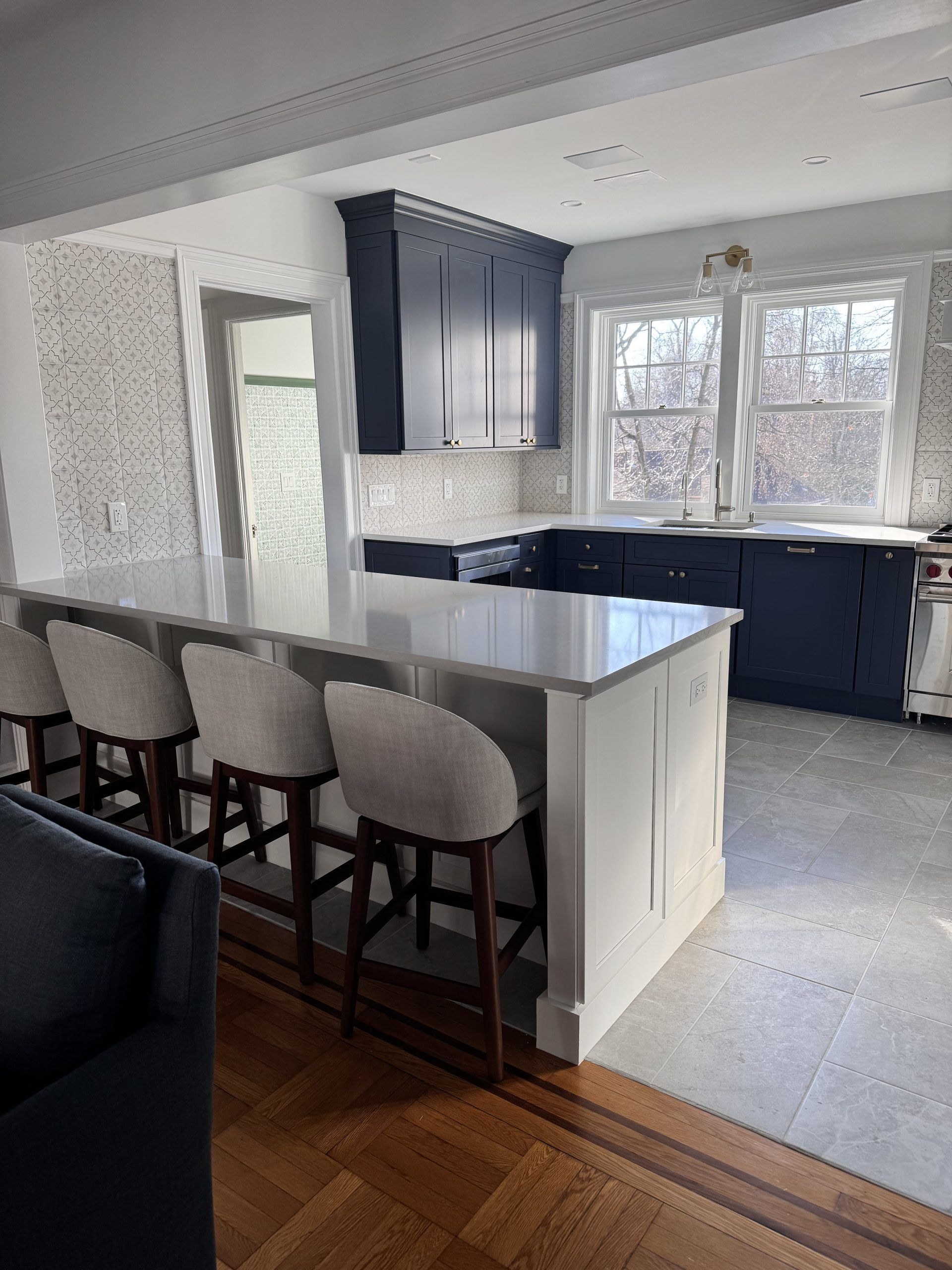 Bright kitchen with white island, three barstools, dark cabinets, and large windows.