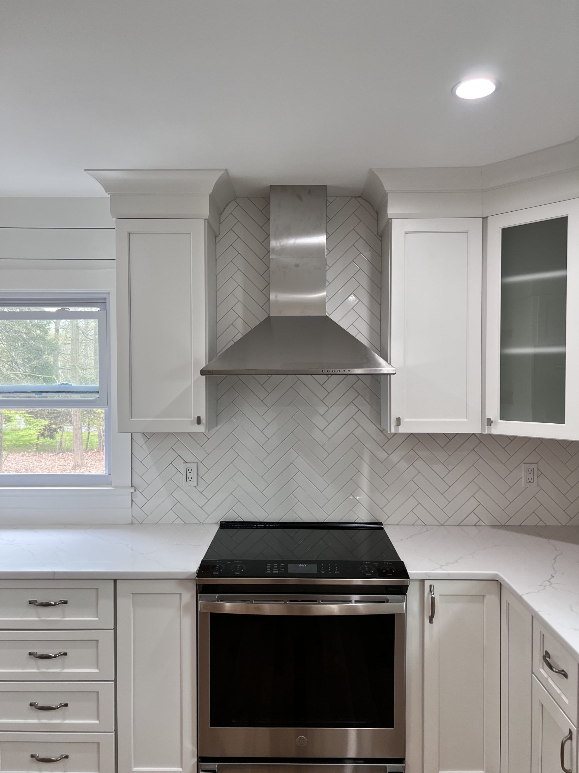 Modern white kitchen with stainless steel range hood, herringbone tile backsplash, and stove under a window