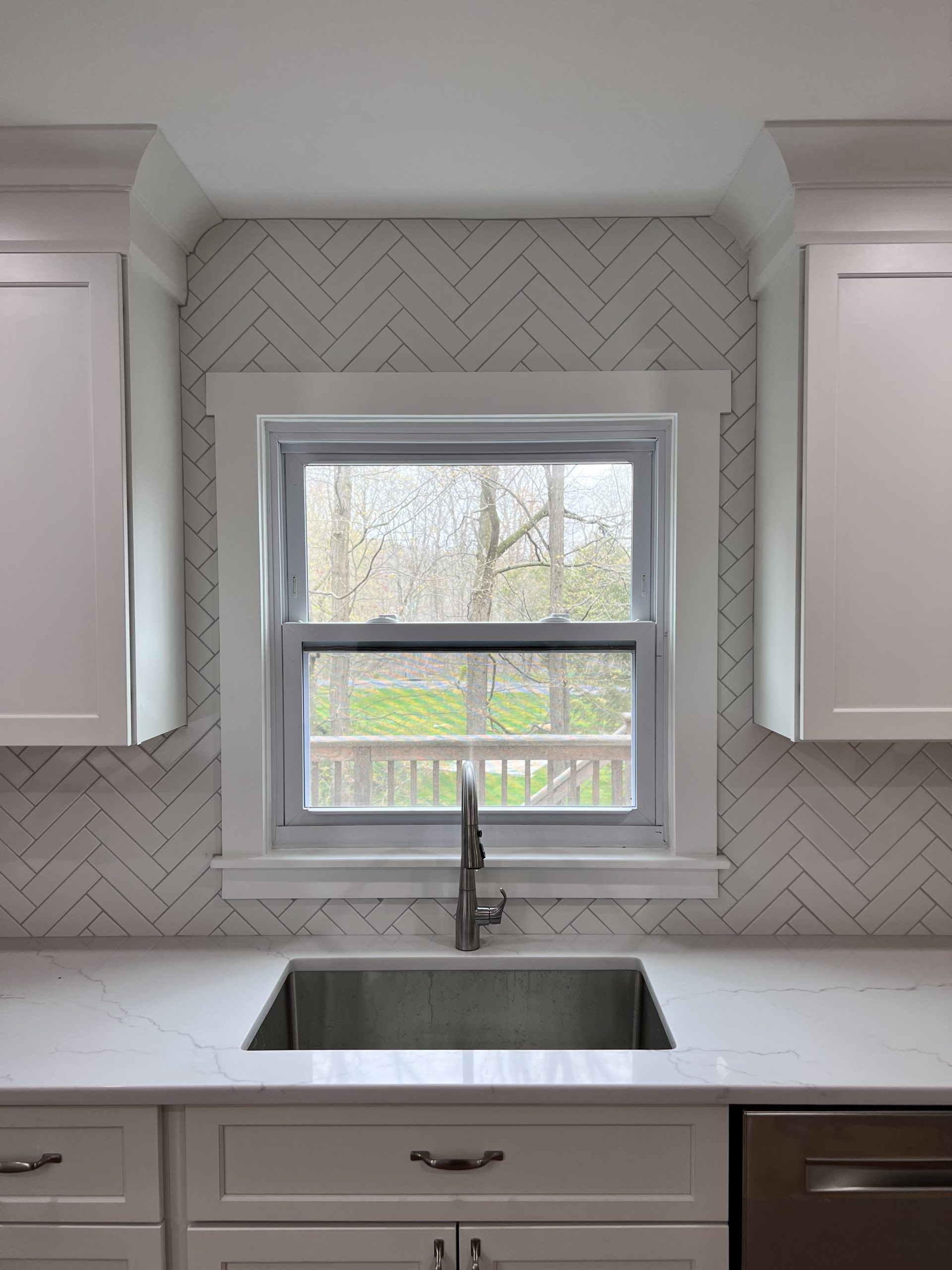 Bright kitchen sink beneath a window with chevron tile backsplash and white cabinets