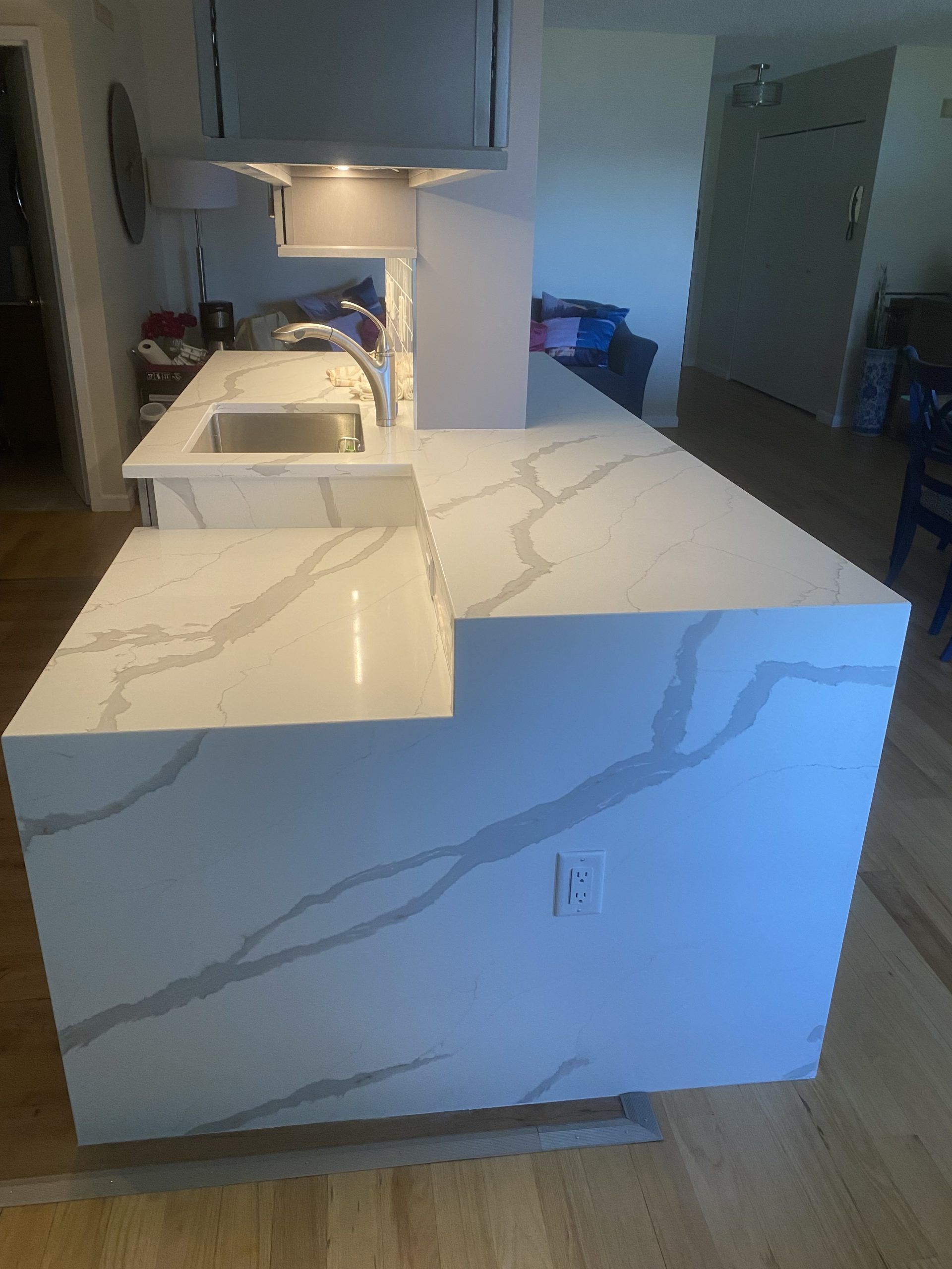 White marble kitchen island and counters in a modern home, with wood floors and a sink under cabinet lighting.
