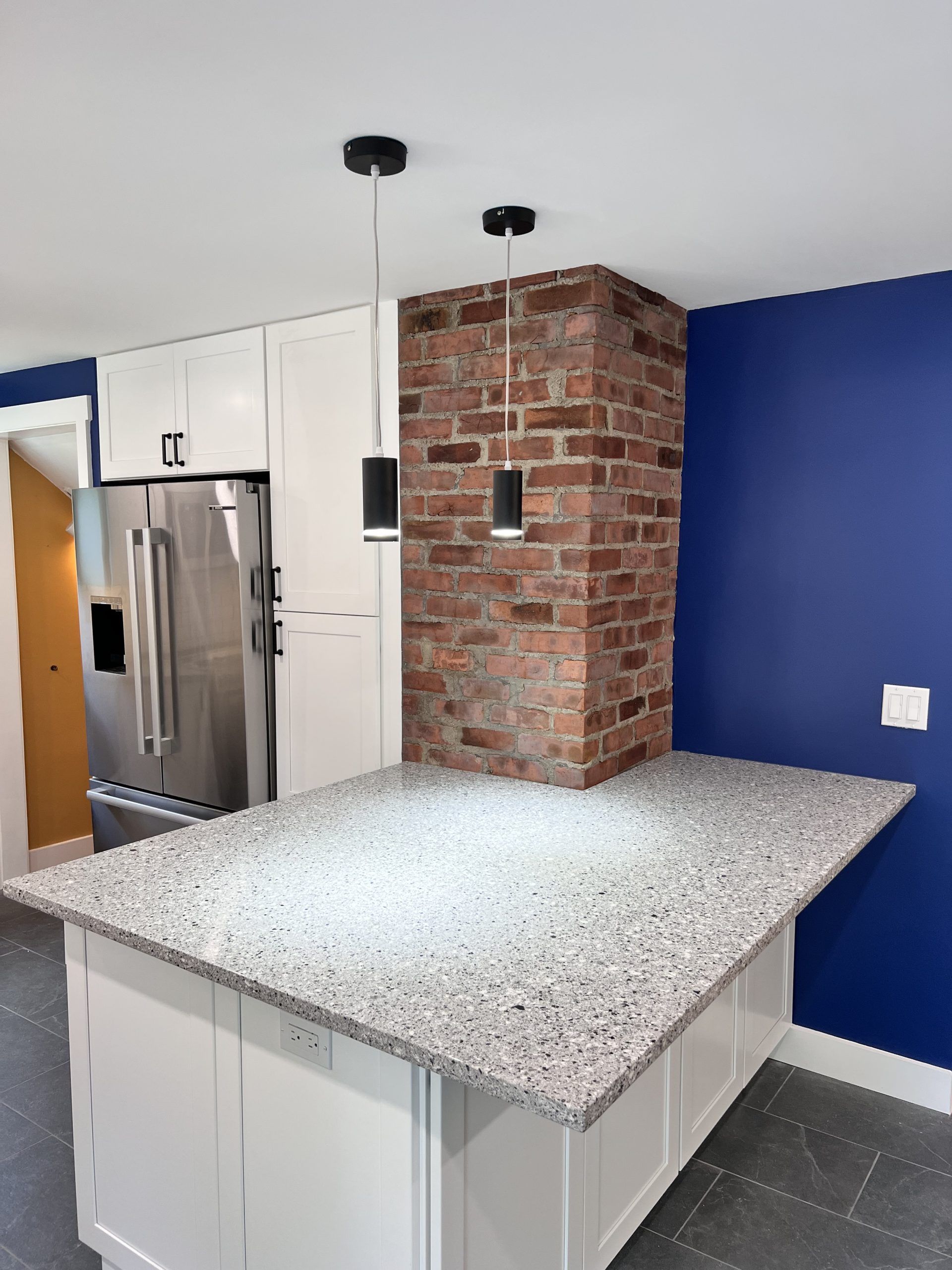 Modern kitchen island with speckled countertop, stainless fridge, white cabinets, brick column, and blue walls