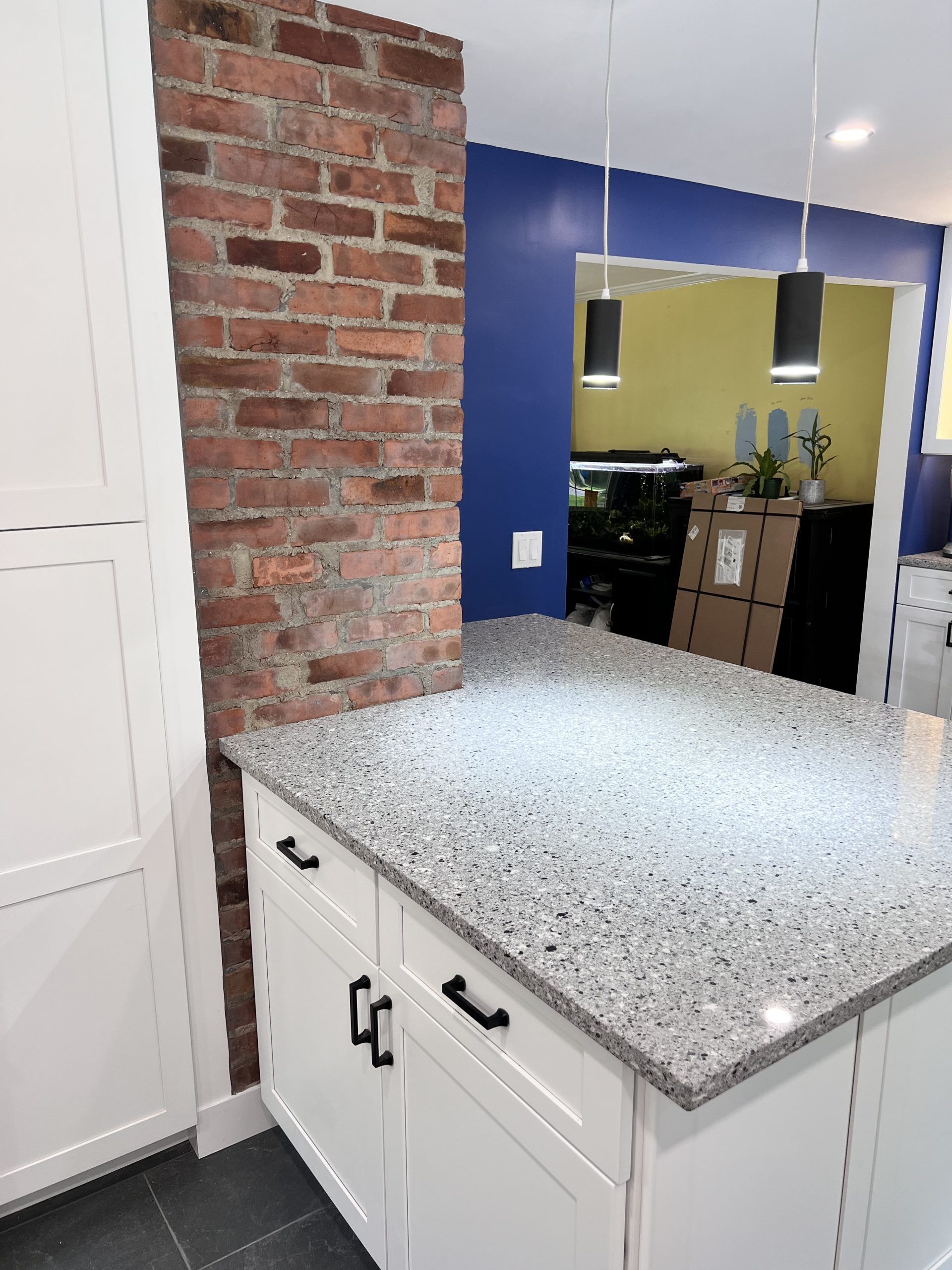 Kitchen island with speckled countertop, white cabinets, exposed brick wall, and blue accent wall.