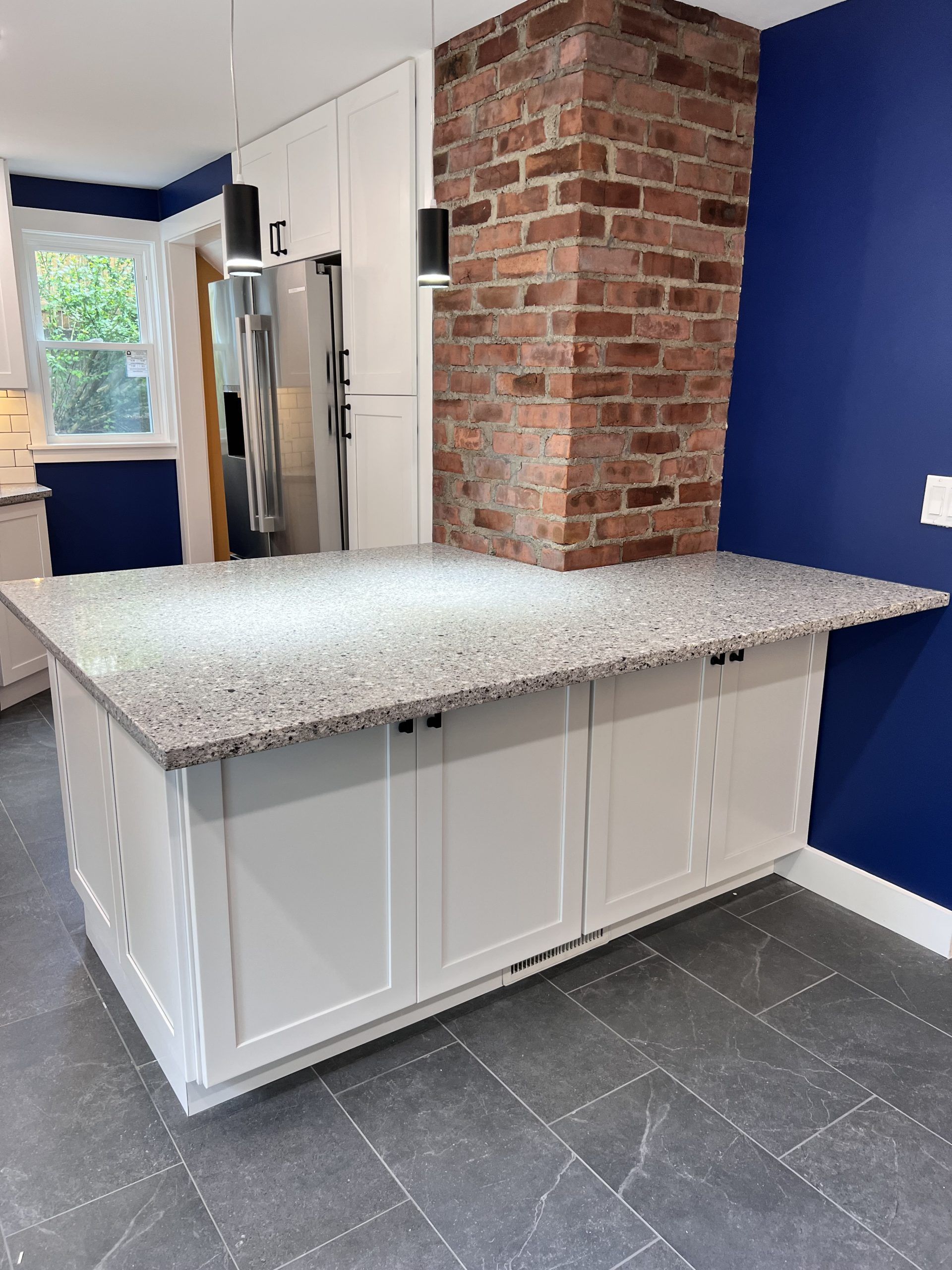 Kitchen island with speckled countertop, white cabinets, exposed brick column, and blue walls