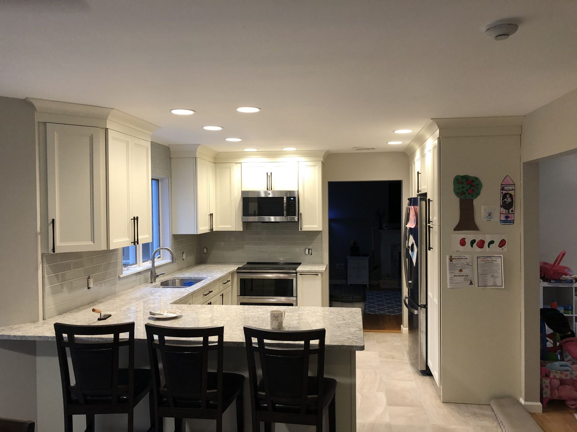 Bright white kitchen with island, black stools, stainless appliances, and a doorway to a dim hallway.