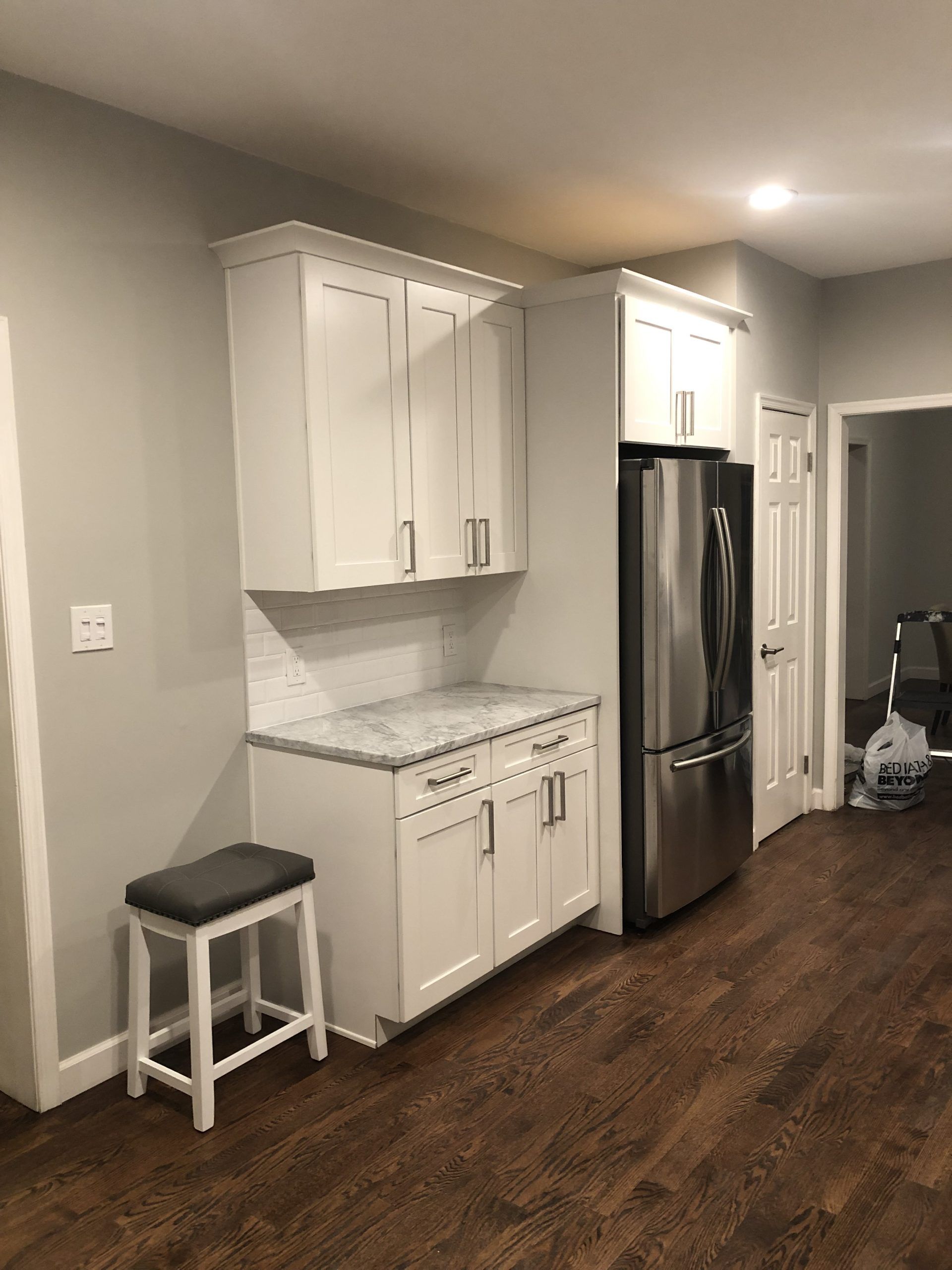 Modern kitchen with white cabinets, stainless steel fridge, dark wood floor, and a small stool by the counter.