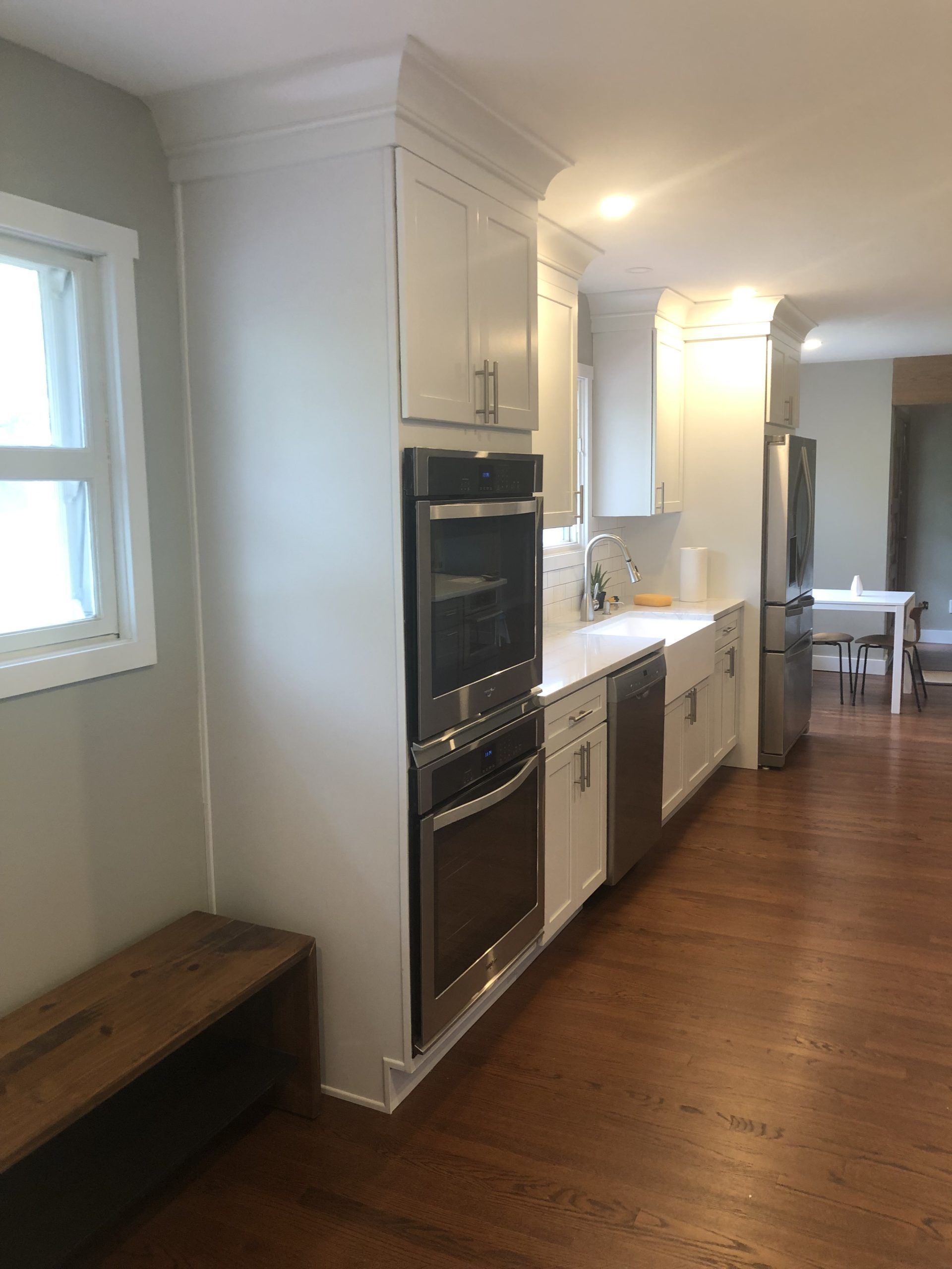 Bright kitchen hallway with white cabinets, wood floors, built-in ovens, and a bench by a window