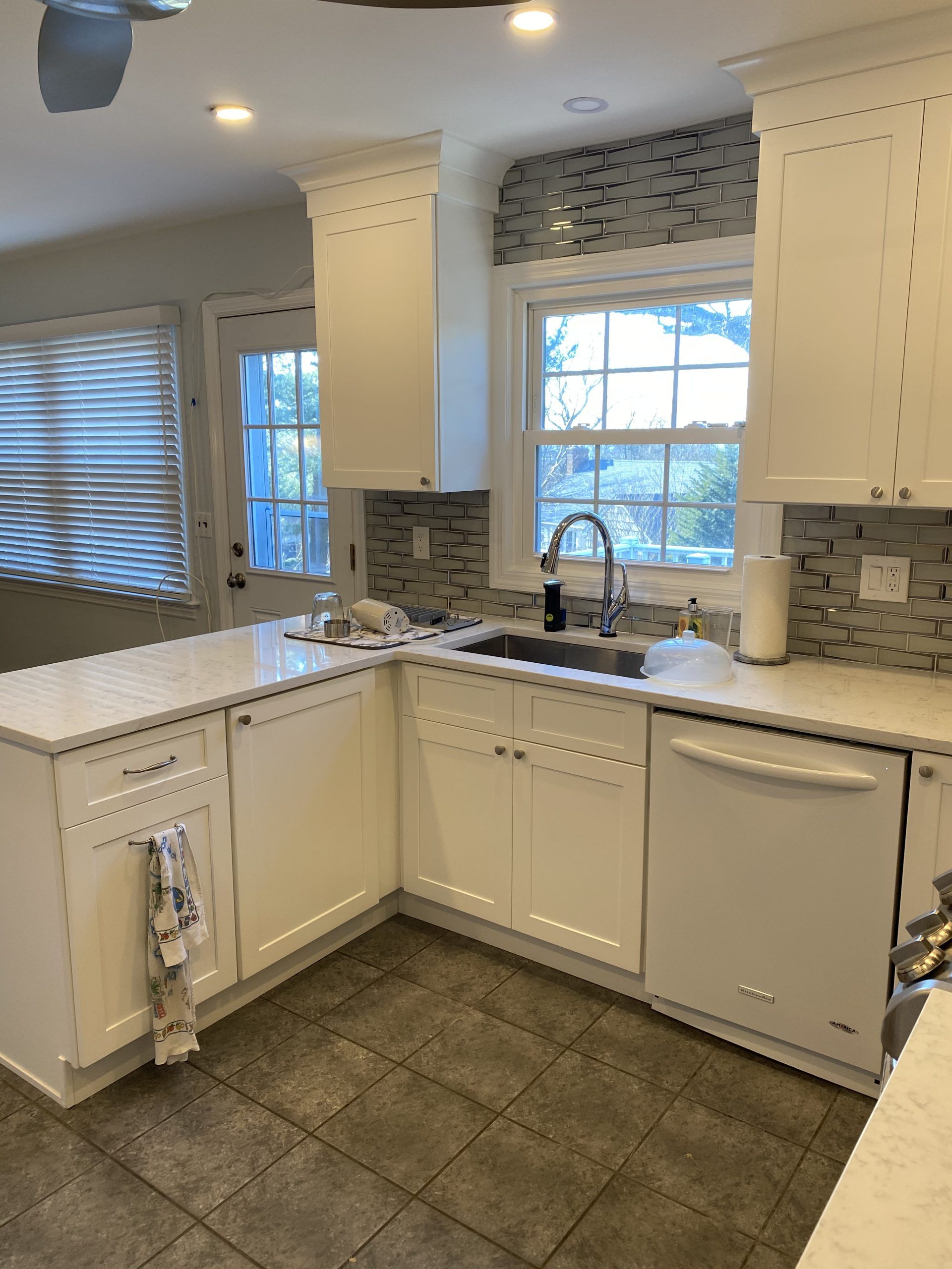 Bright kitchen with white cabinets, gray tile backsplash, window over sink, and dark tiled floor.