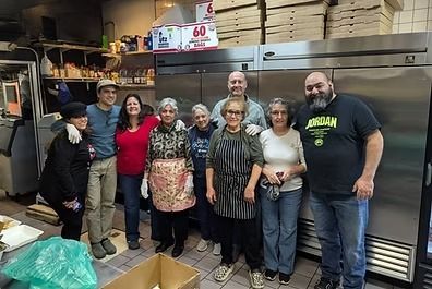 Group of people in a commercial kitchen; some wearing aprons. They are smiling and posing near large refrigerators.