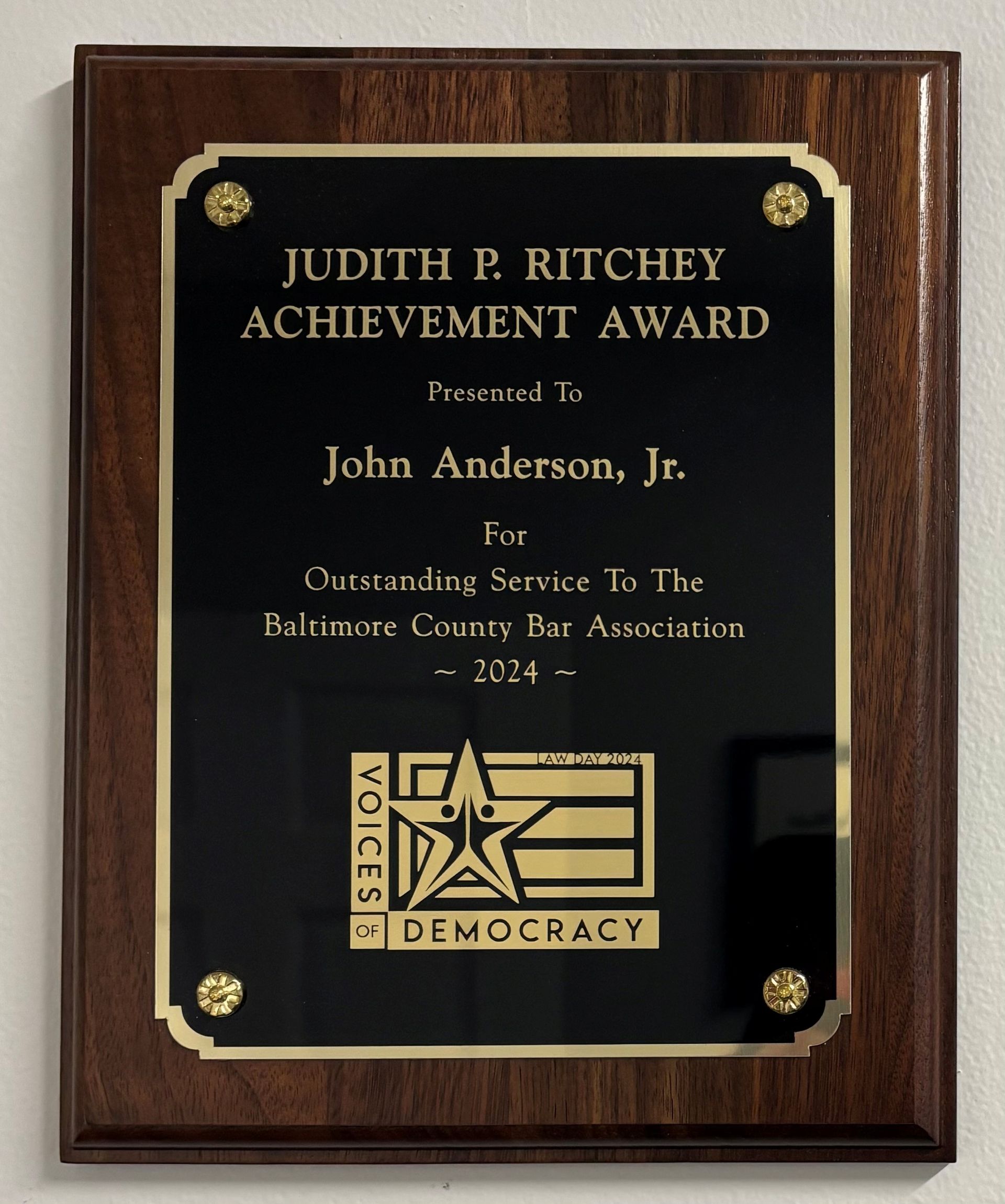 Woman and man holding an achievement award. Man wears a suit, woman wears a black top. Background: framed artwork.