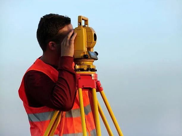 Surveyor in orange vest using a theodolite on a tripod, outdoors against a blue sky.